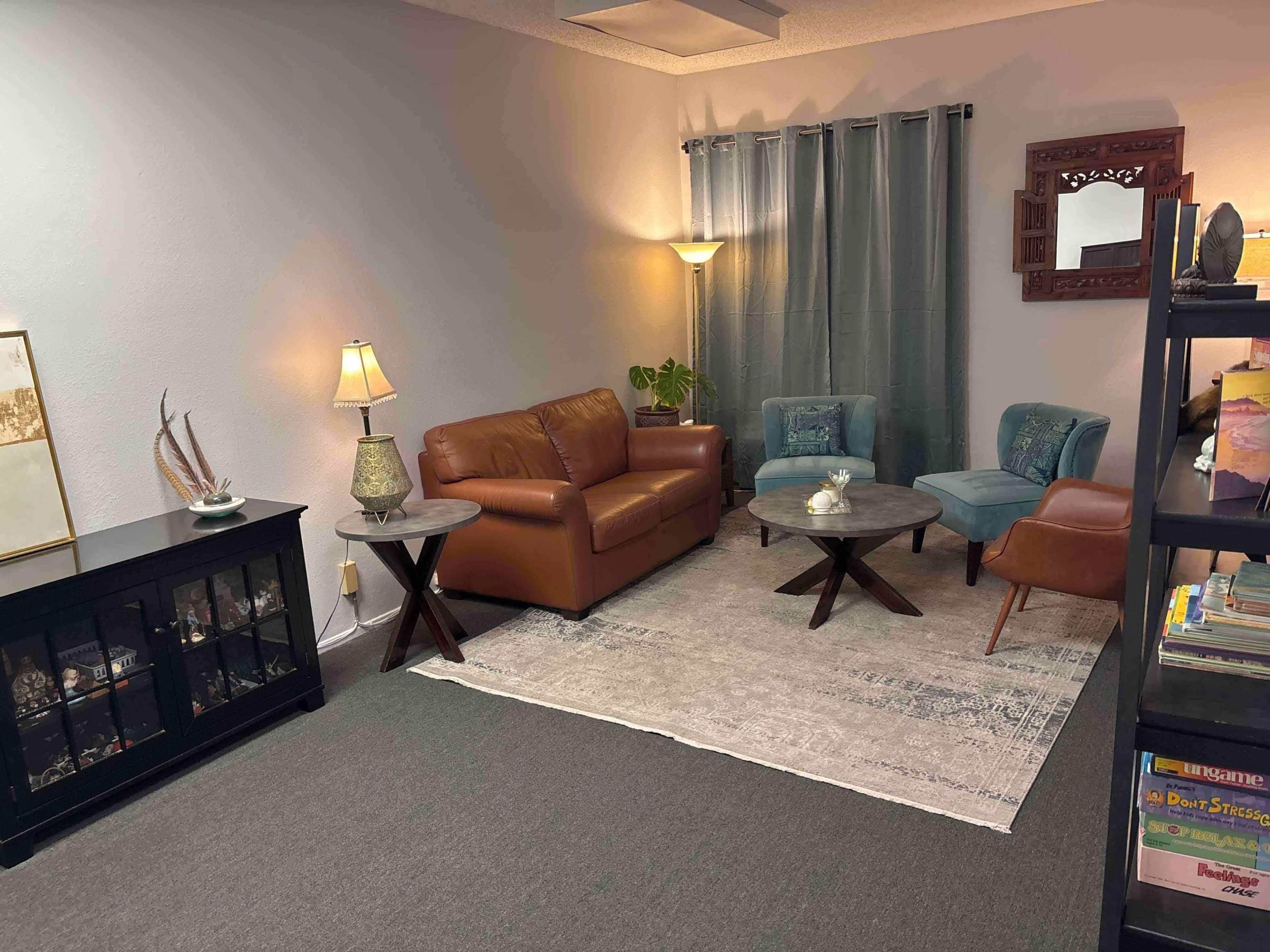 The image shows a cozy living room with a brown leather sofa, two blue armchairs, a couple of coffee tables, and a light source from a floor lamp, all arranged on a light-colored rug.