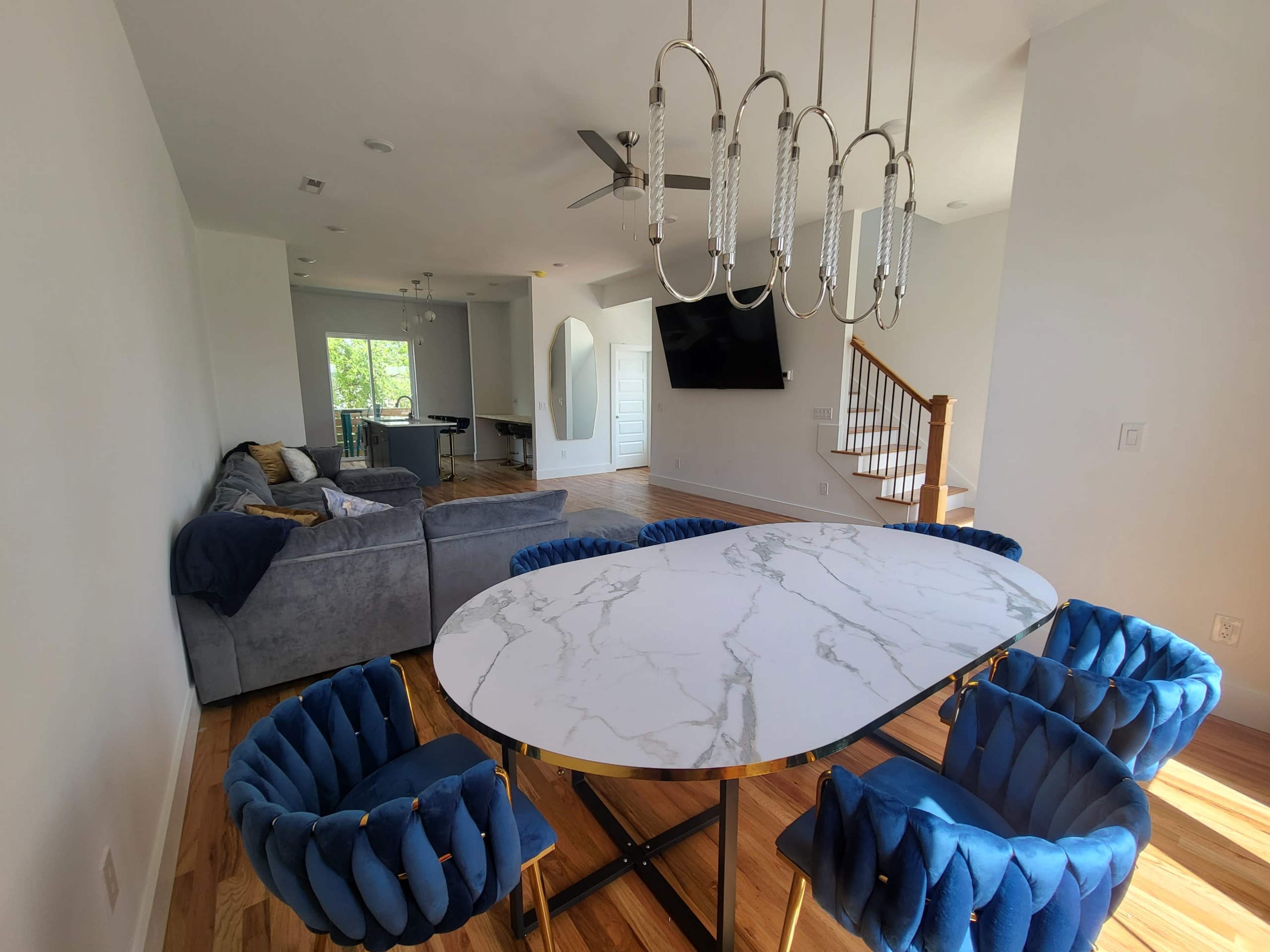 A modern dining area features a marble-top oval table surrounded by blue velvet chairs, with a spacious living room visible in the background.