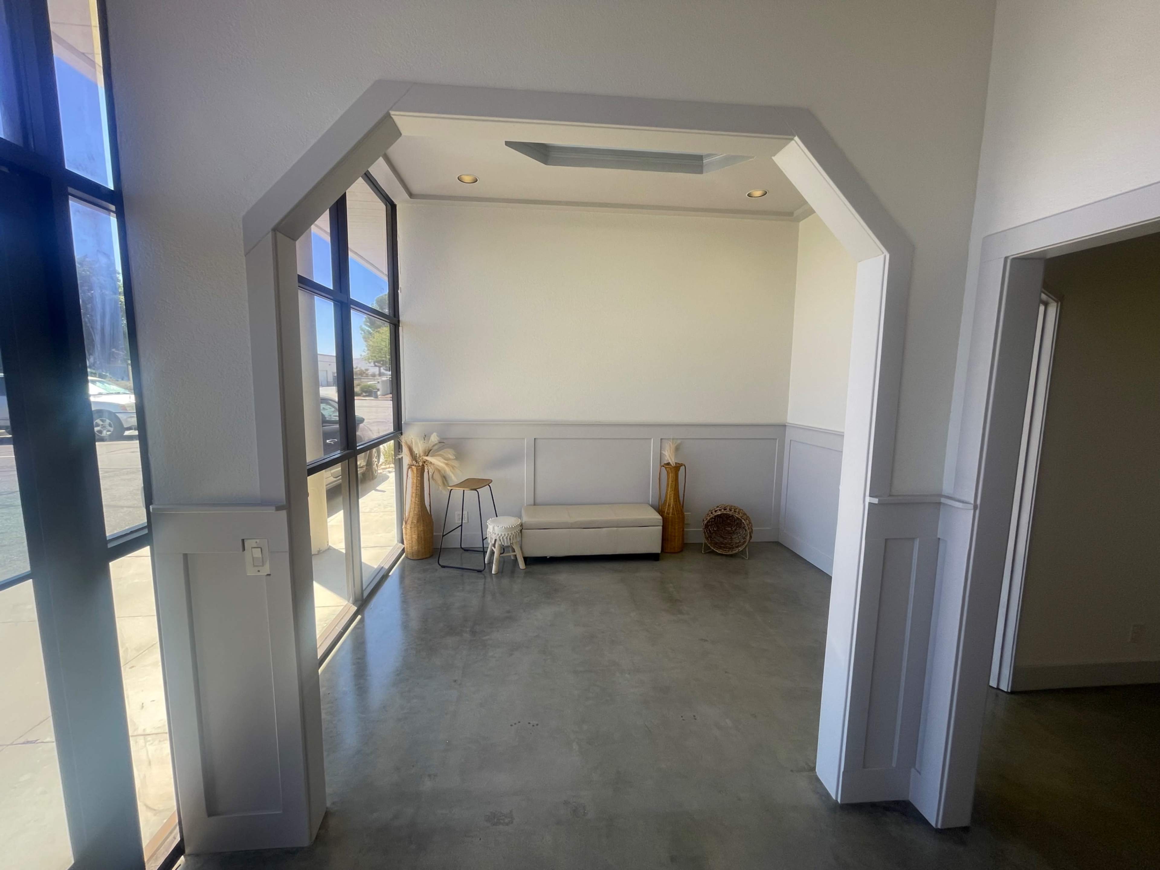 The image shows a minimalist interior space framed by an arched entryway, featuring a light-colored bench, a small stool, and decorative vases near large windows.