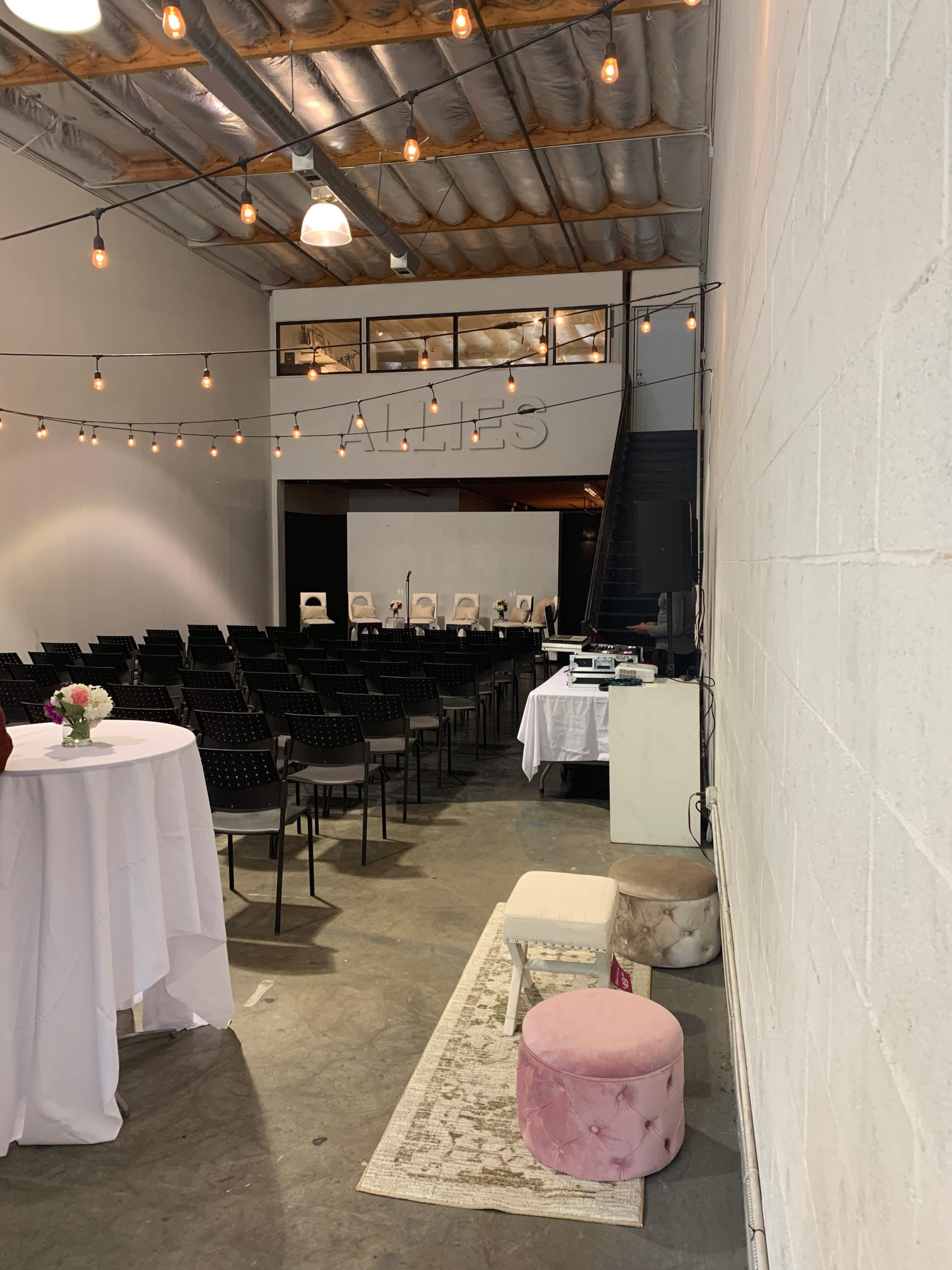 A spacious indoor event venue with rows of black chairs, decorative string lights overhead, and a small seating area with pastel stools and a table in the foreground.