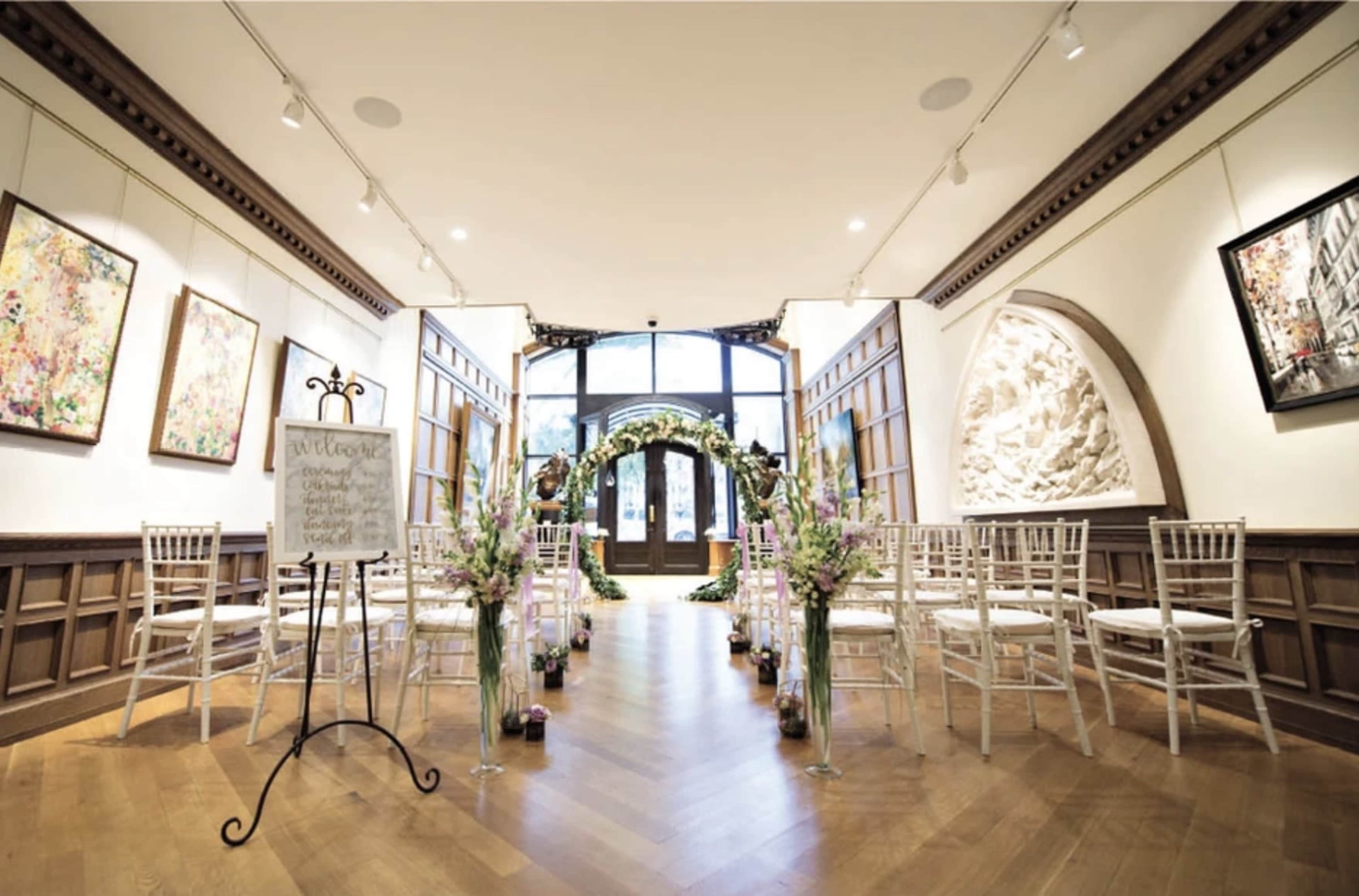 A wedding ceremony setup features rows of white chairs facing an ornate arch adorned with greenery and flowers in a tastefully decorated gallery.