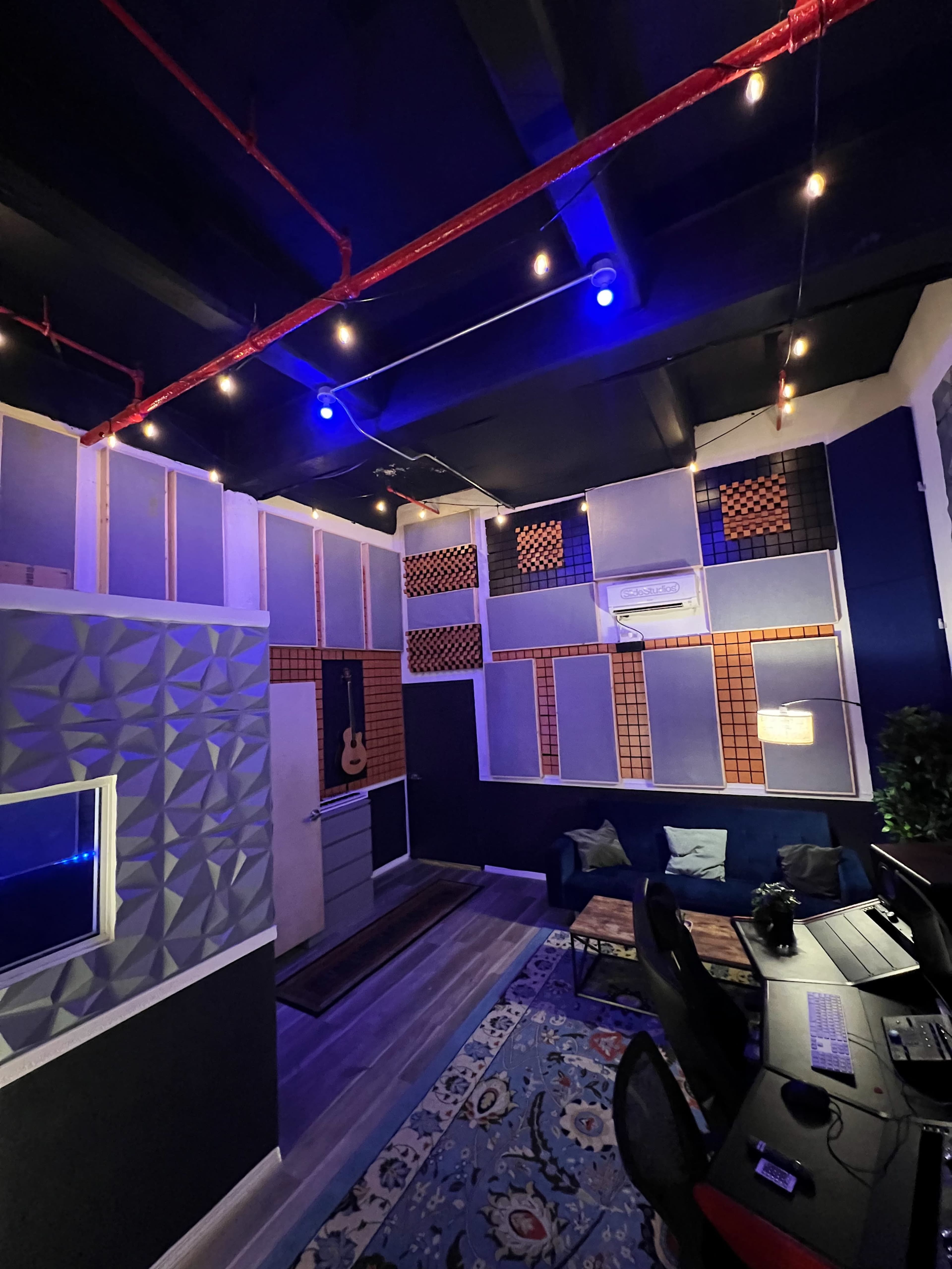 A soundproof recording studio with acoustic panels, a guitar hanging on the wall, and a seating area with a coffee table and computers.