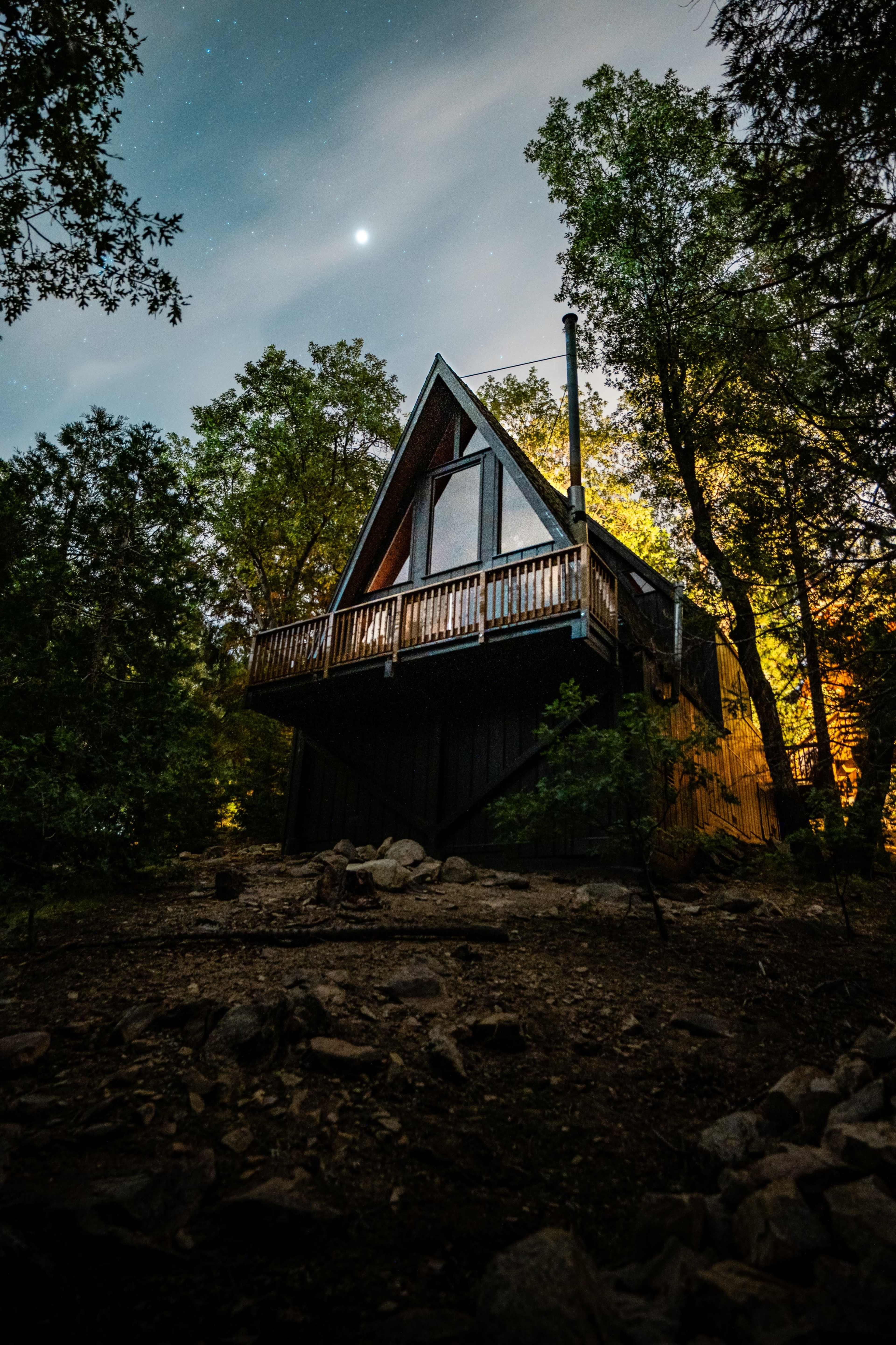 A triangular-roofed cabin stands on stilts amid trees under a starry sky.