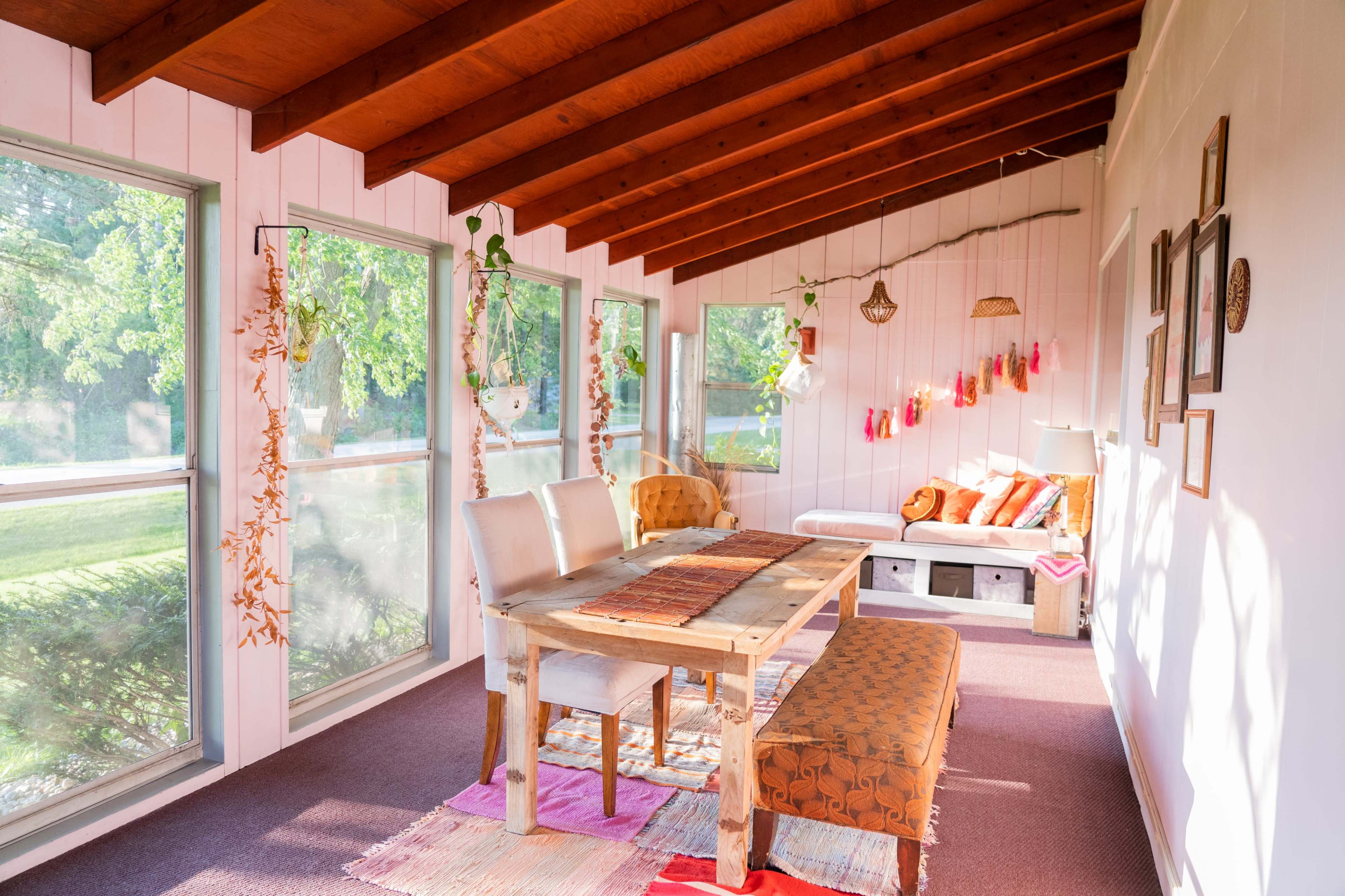 A sunlit enclosed porch features a wooden dining table with white chairs, a comfortable seating area adorned with cushions, and large windows overlooking greenery.