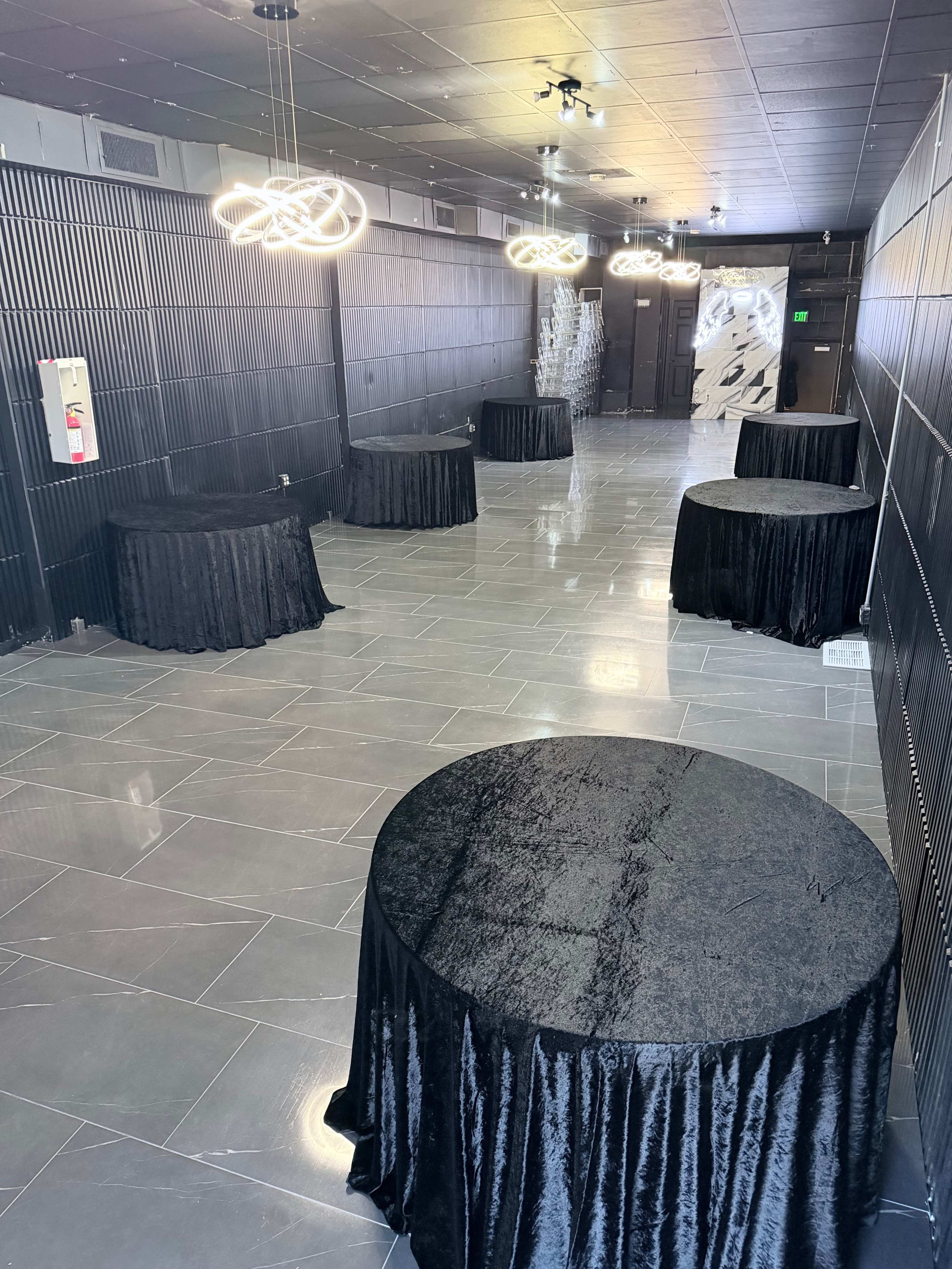 The image shows a spacious, modern room featuring several round tables covered with black tablecloths under contemporary light fixtures.