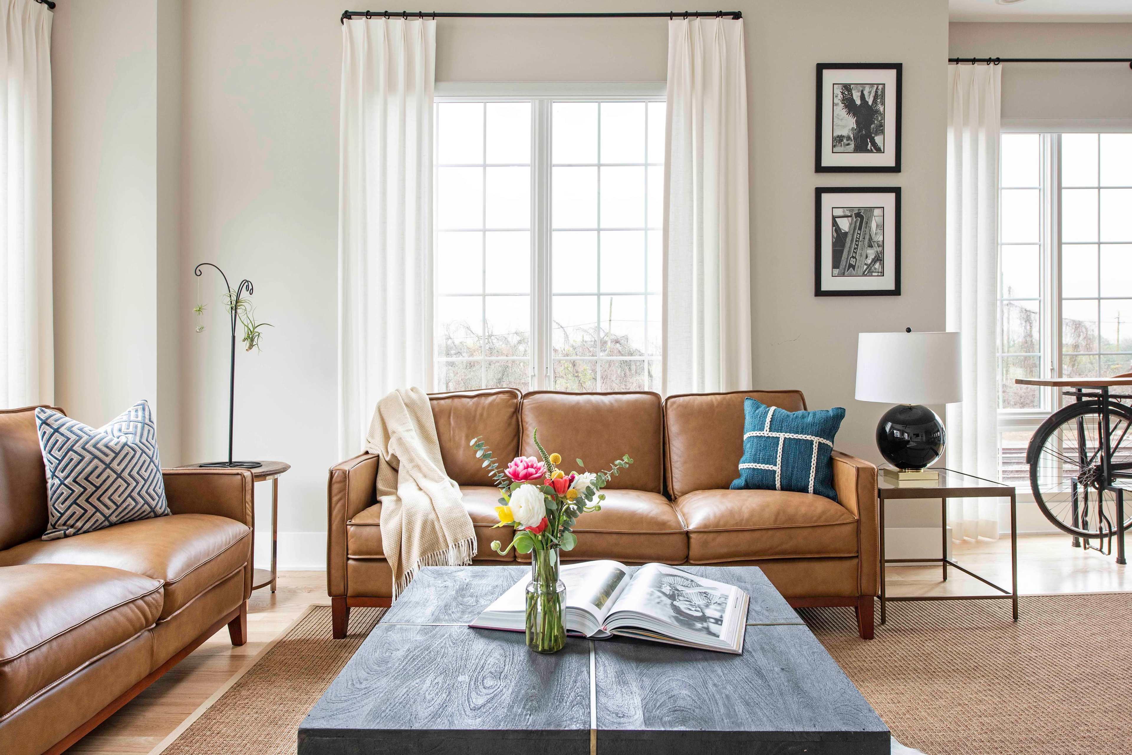 The image shows a modern living room with two leather sofas, a coffee table on a rug, and large windows featuring light curtains.