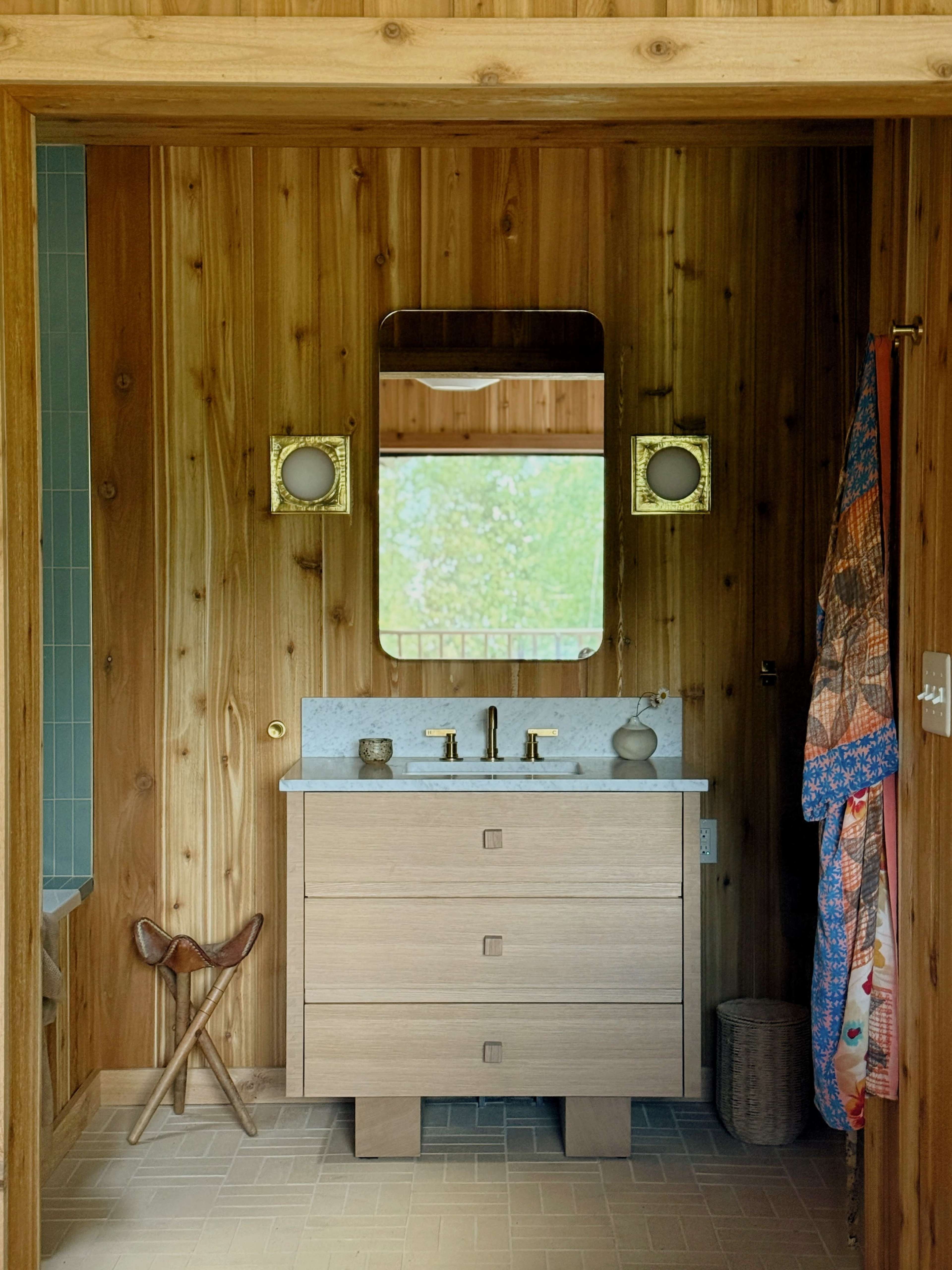 A bathroom features a wooden wall, a marble-topped vanity with gold faucets, a large mirror, and a colorful towel hanging nearby.