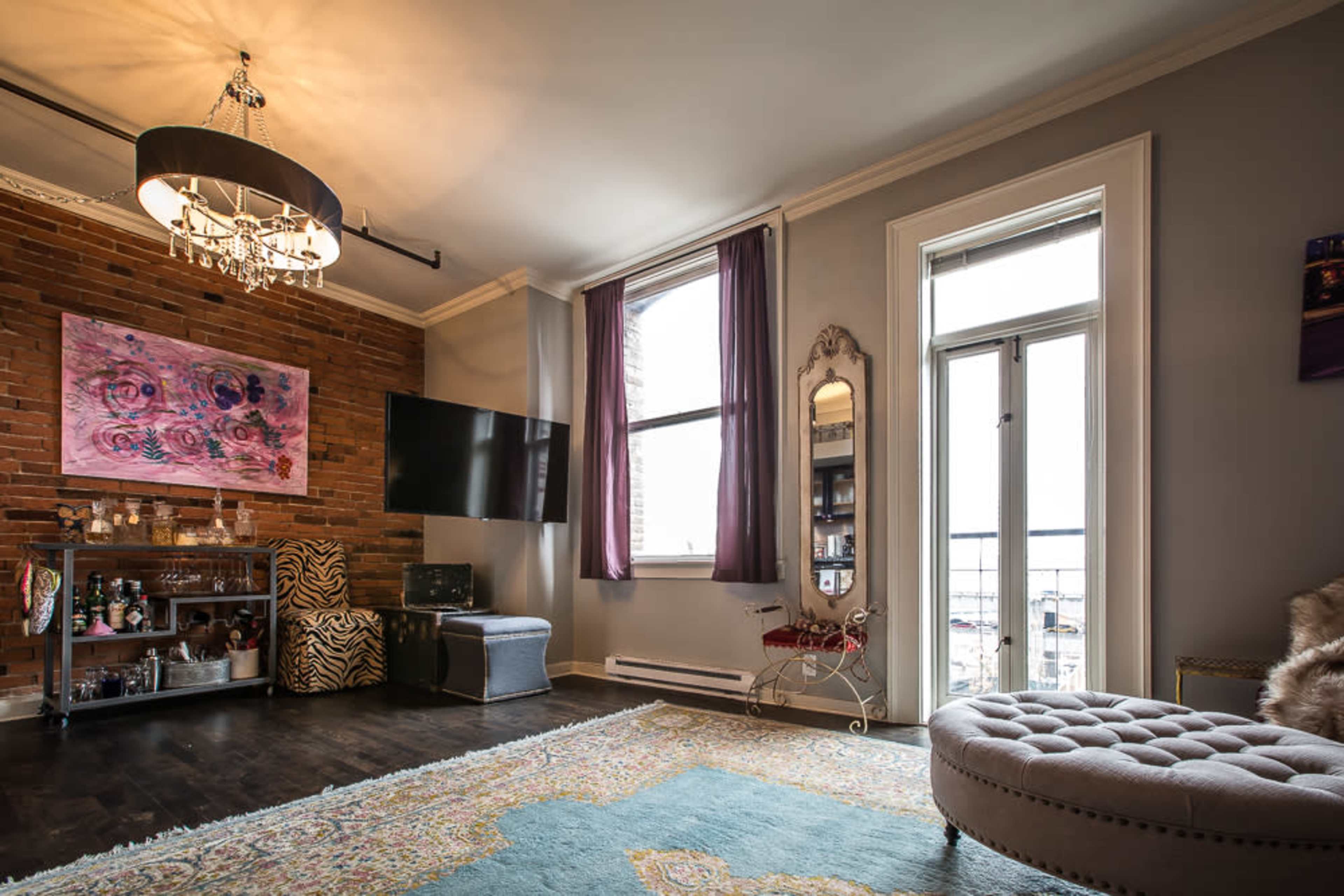 The image shows a living room with a sectional sofa, a wall-mounted television, a decorative mirror, and a large window with curtains, complemented by a colorful rug and artwork on the walls.