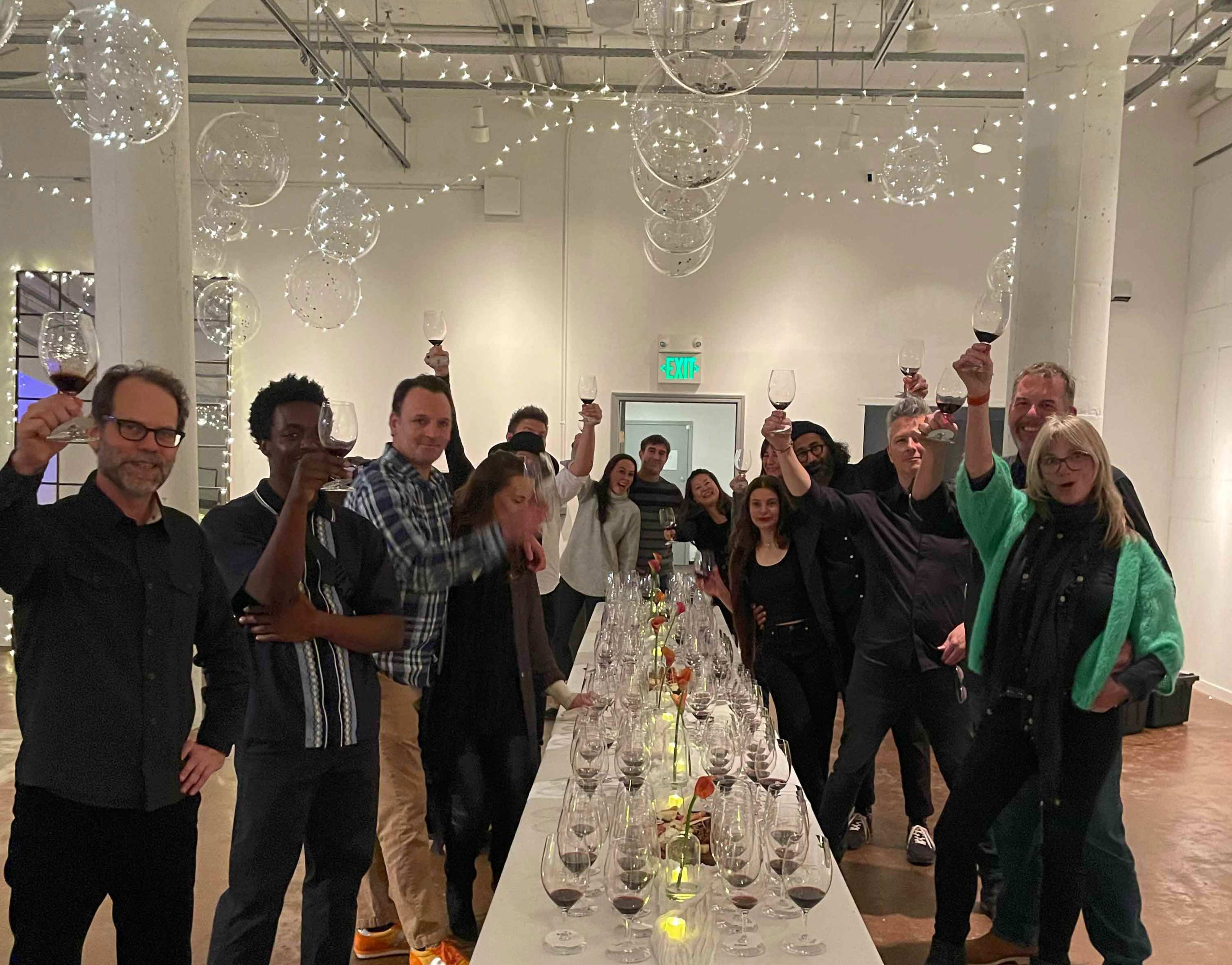 A group of people are toasting with wine glasses around a long table decorated with lights and glass ornaments in a brightly lit room.