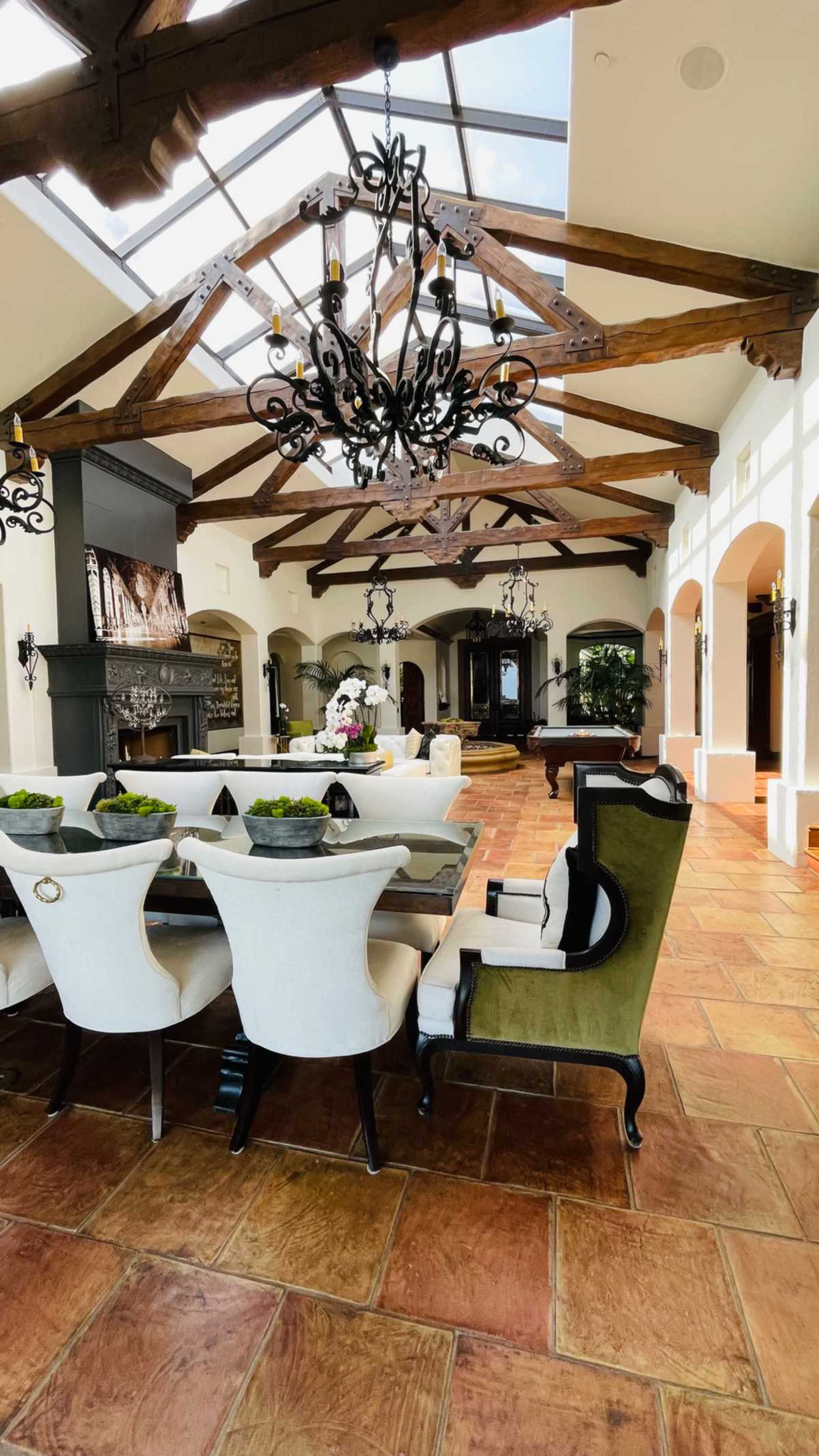 The image shows a spacious dining area featuring a long table surrounded by upholstered chairs, a large chandelier, and exposed wooden beams in the ceiling.