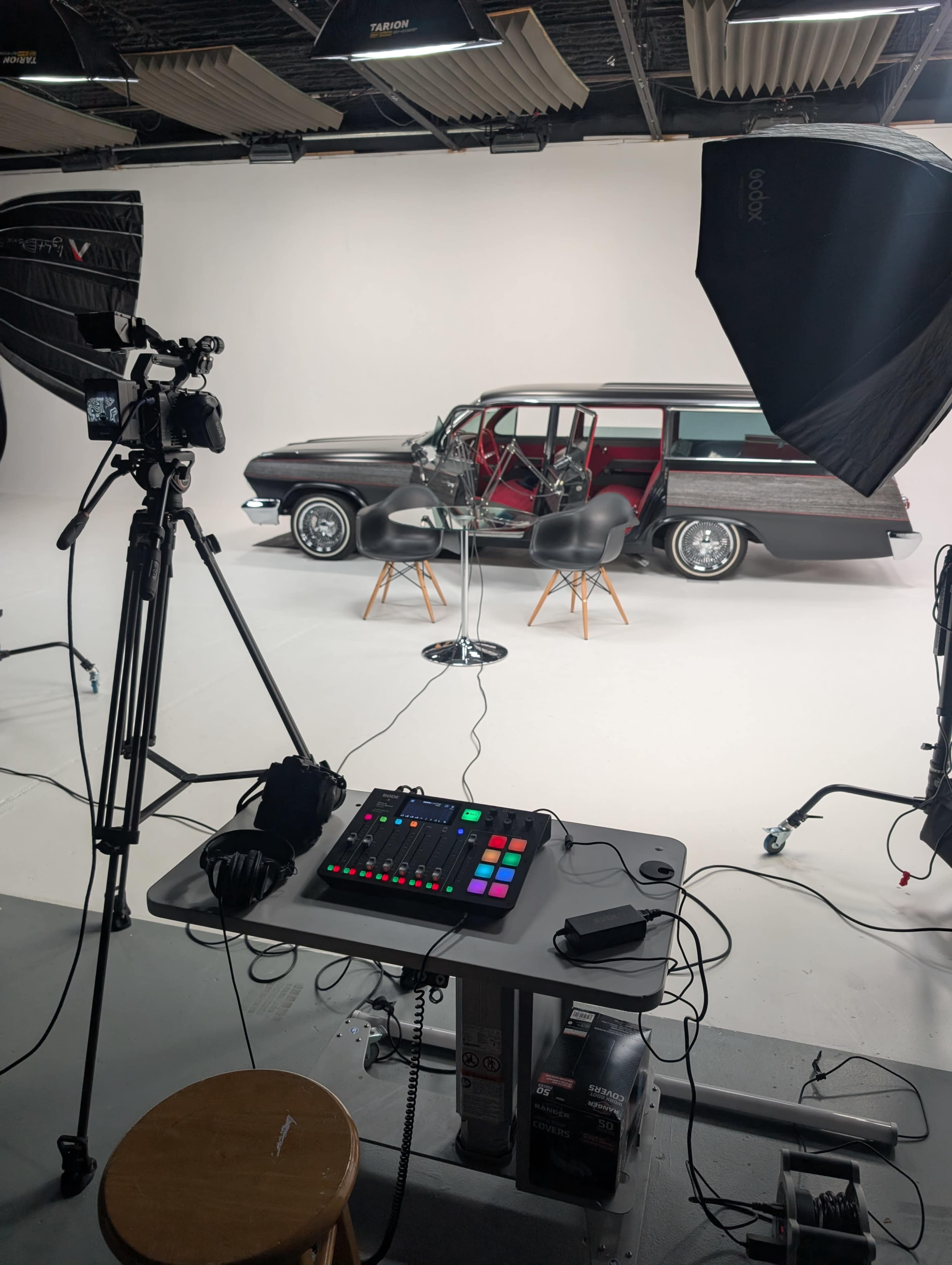 The image shows a photography studio setup with a vintage car in the background, two chairs facing each other, and various lighting and camera equipment in the foreground.