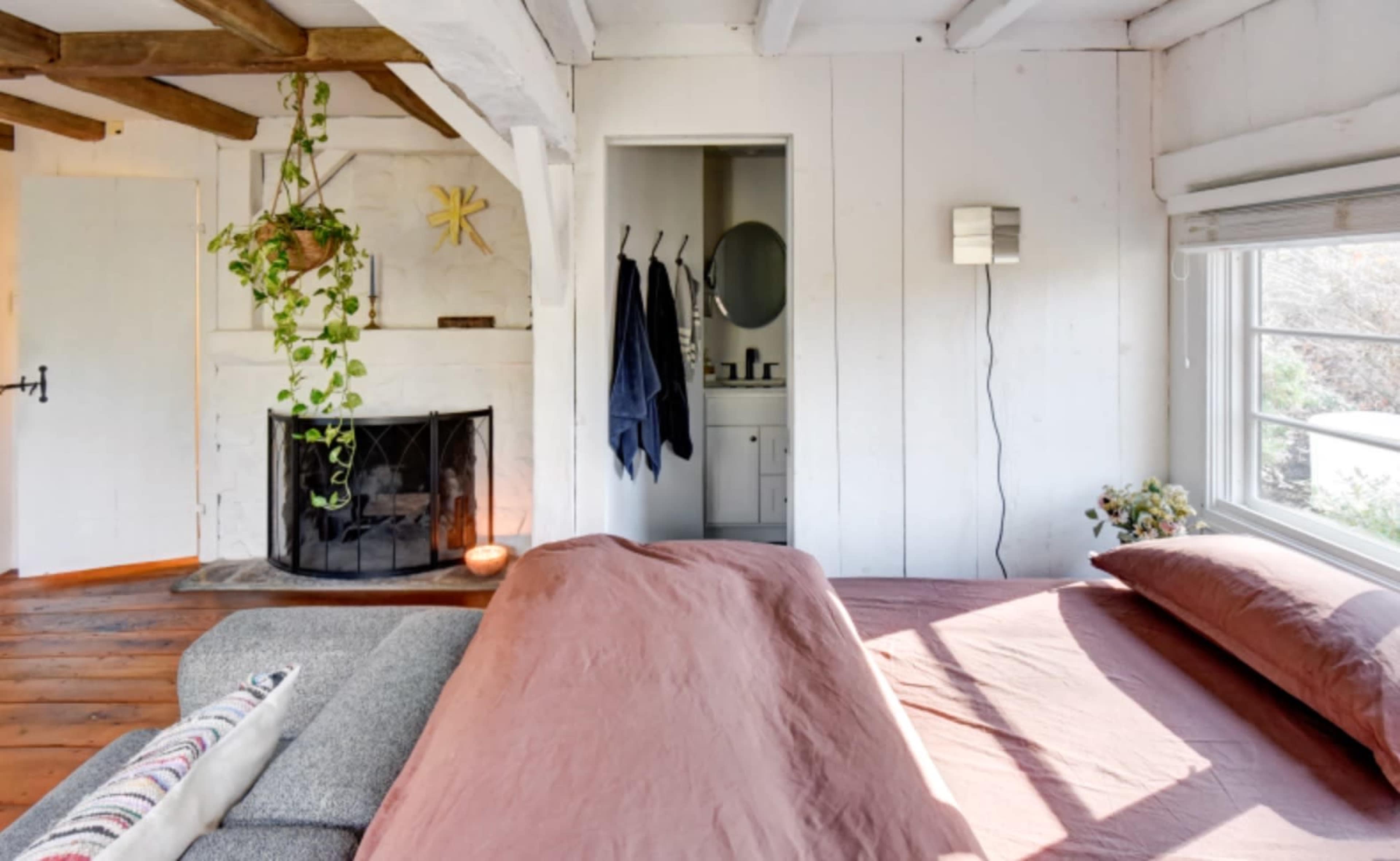 A cozy bedroom features a bed with earthy-colored linens, a fireplace, and an adjoining bathroom visible through an open door.