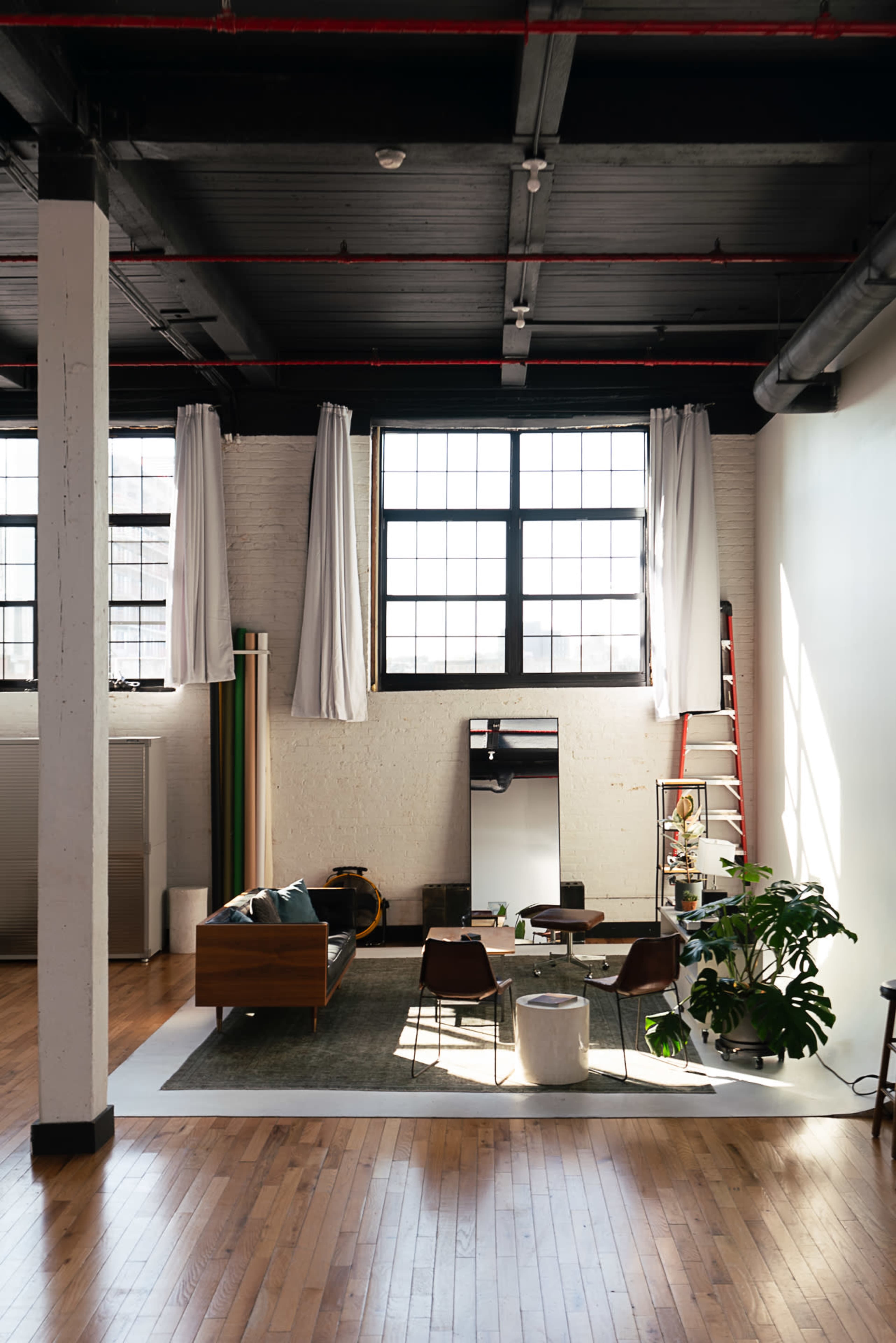 A spacious loft features large windows, wooden floors, and a sitting area with a sofa, chairs, and plants.