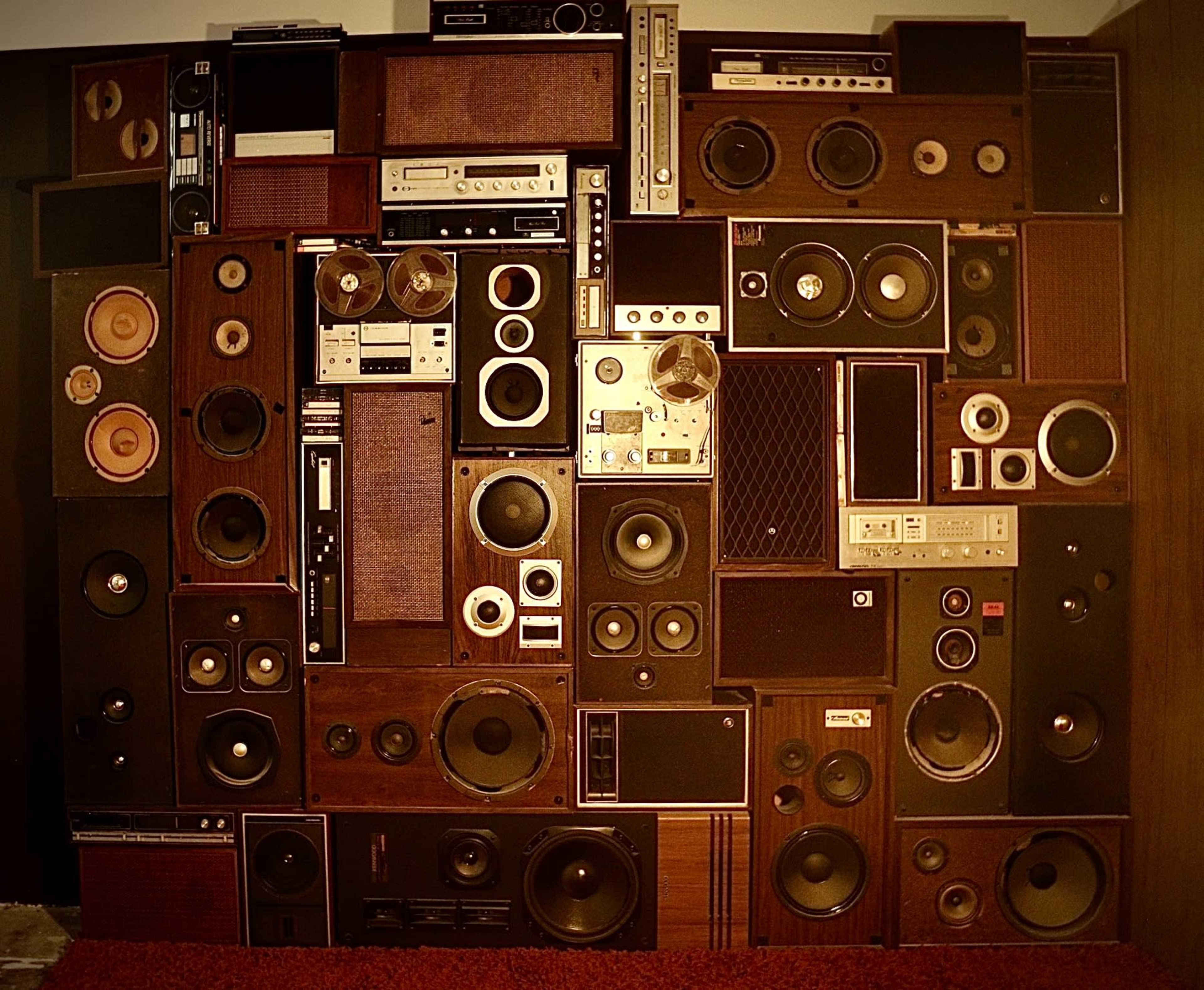 The image shows a wall covered with a collage of various vintage audio equipment, including speakers, cassette decks, and turntables.