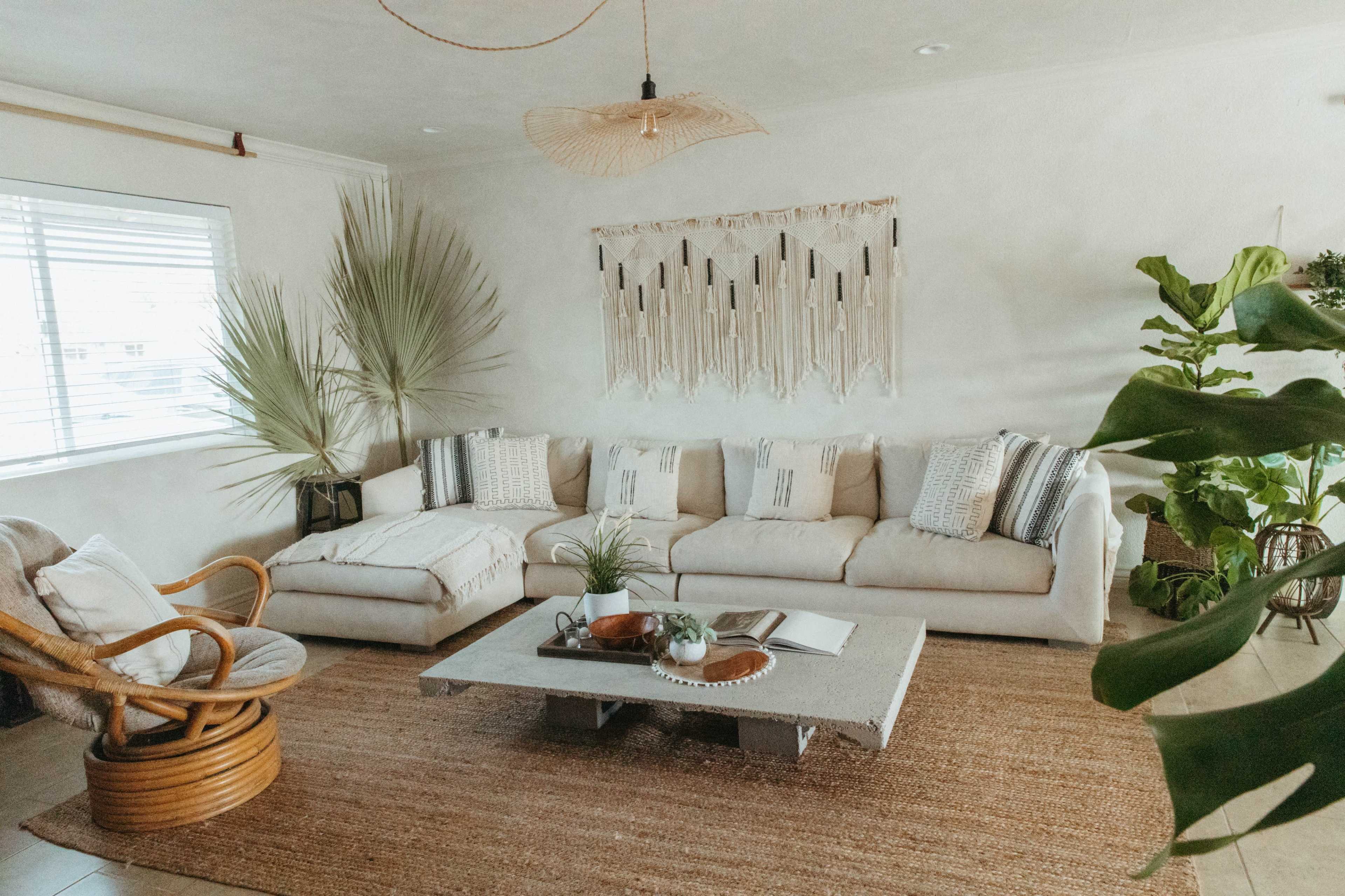 The living room features a large, light-colored sectional sofa, a round rattan chair, and a decorative wall hanging above a coffee table surrounded by plants and a woven rug.