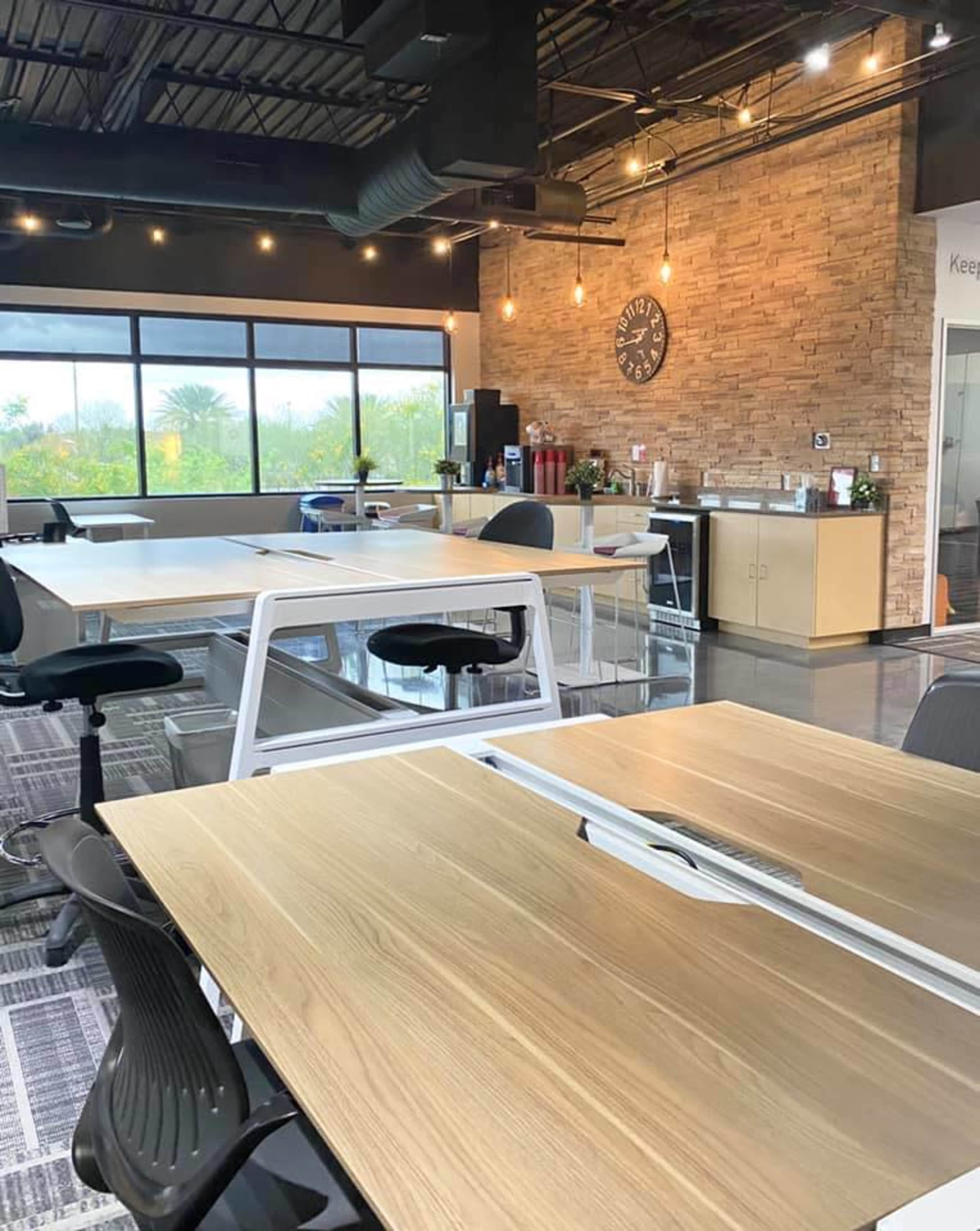 A modern office space with multiple wooden desks, ergonomic chairs, and large windows providing natural light.