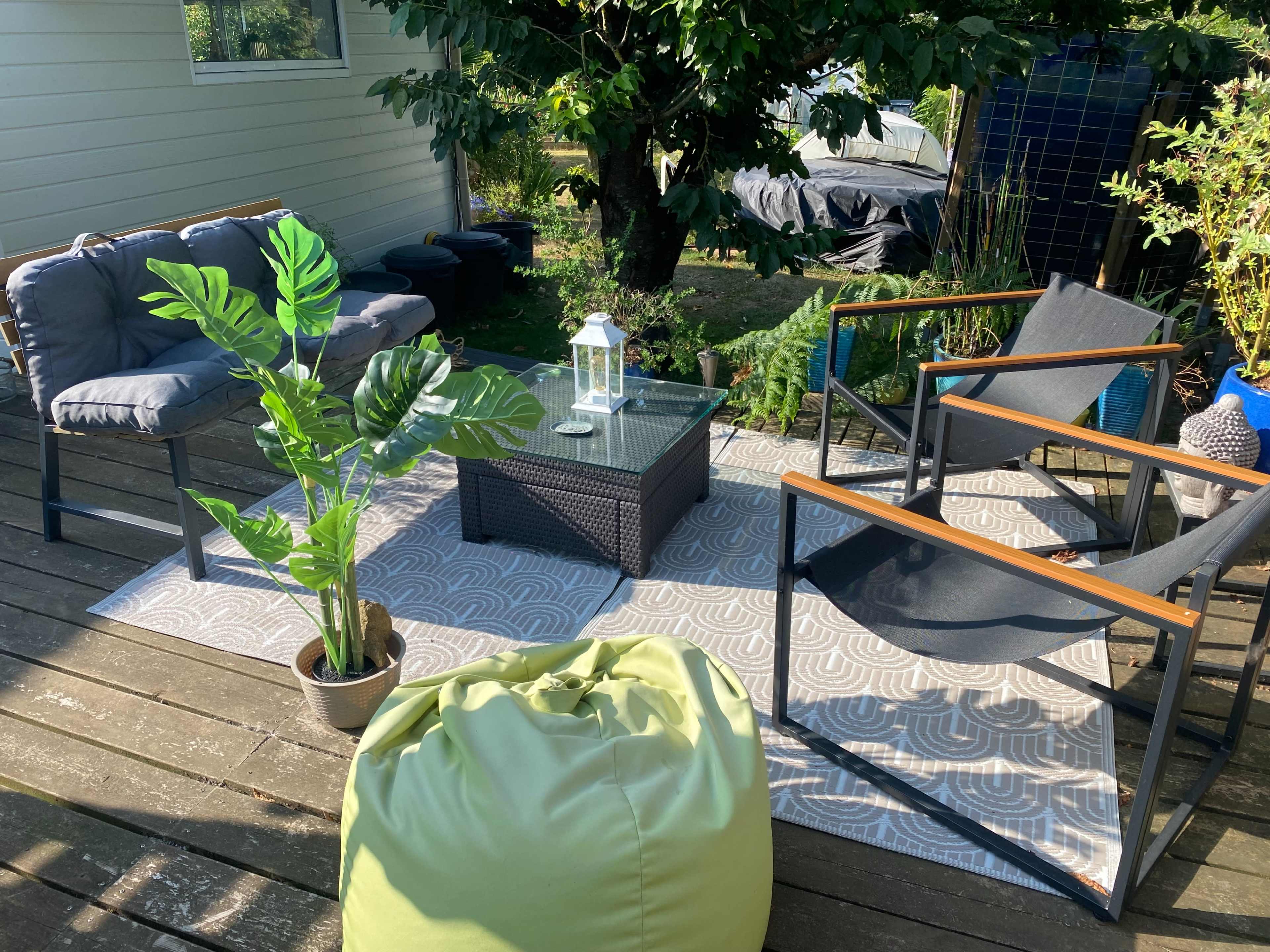 The image shows an outdoor seating area with two armchairs, a couch, a coffee table, a potted plant, and a green bean bag on a patterned rug.