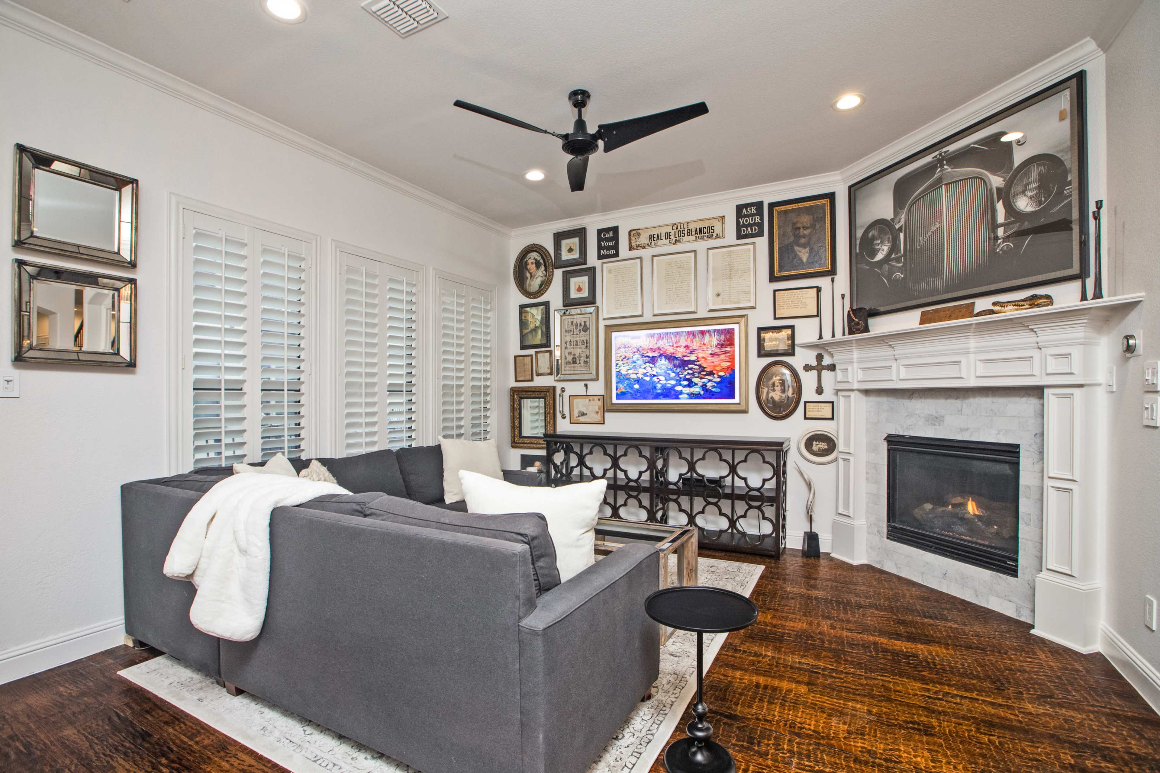 A cozy living room features a gray sectional sofa, a coffee table, and a fireplace surrounded by framed art and photographs on the walls.