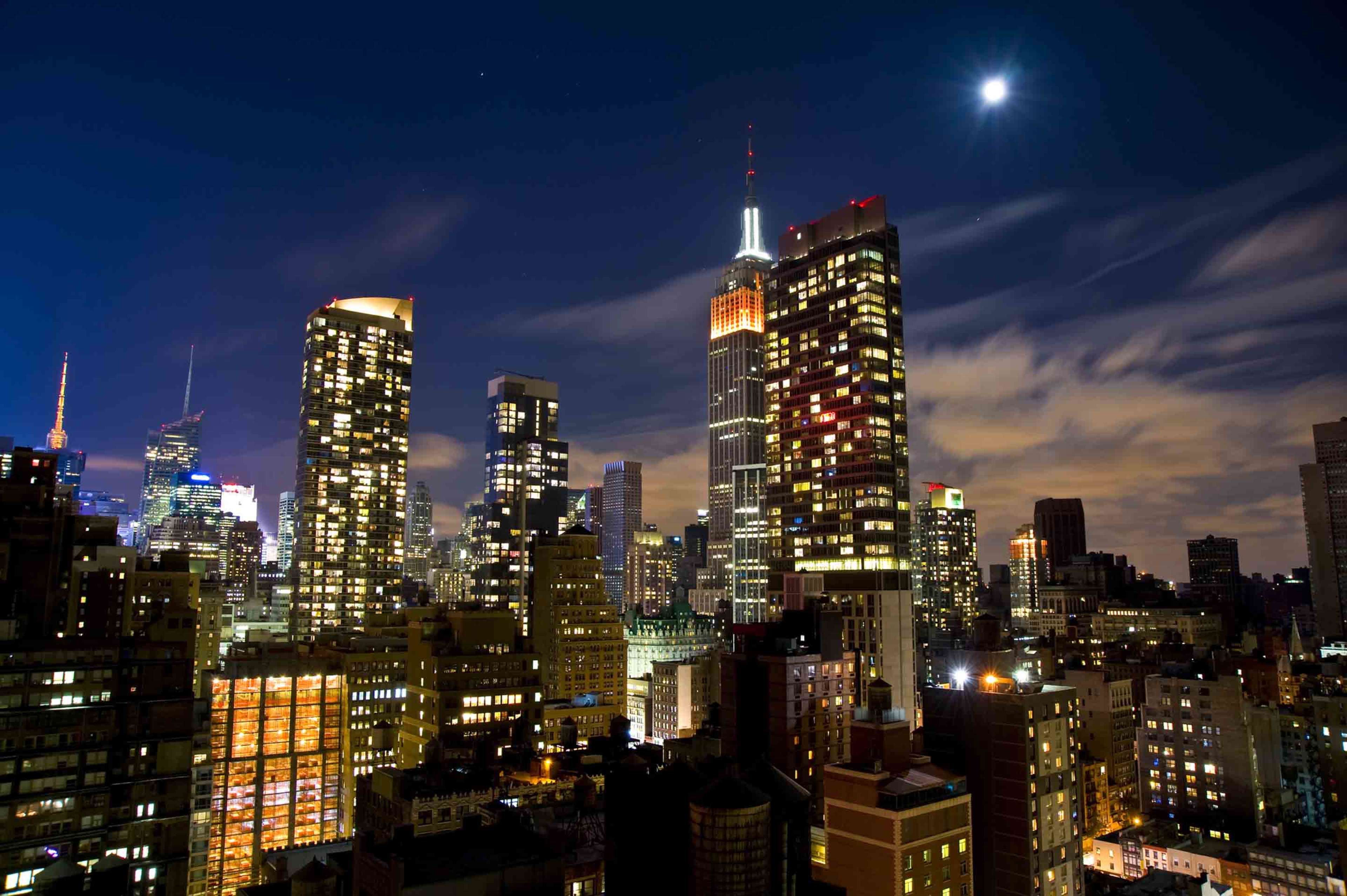 A night cityscape of Manhattan, featuring illuminated skyscrapers and the Empire State Building under a crescent moon.