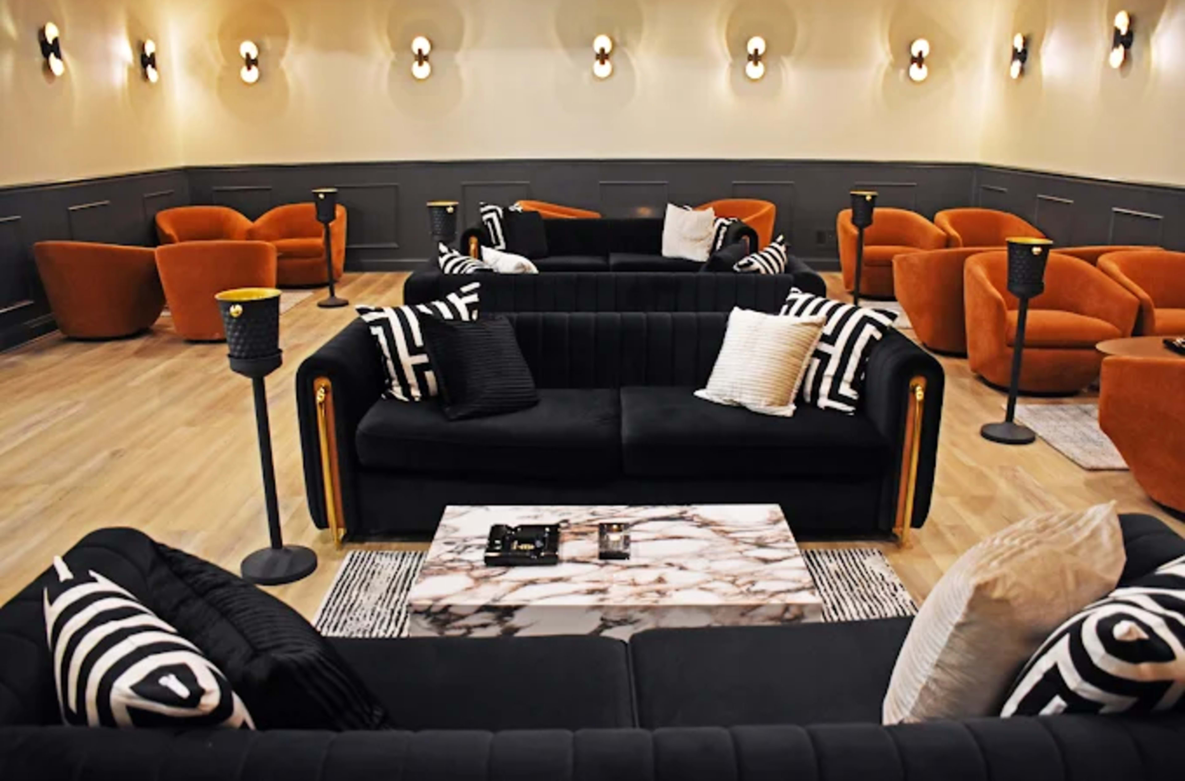 The room features a modern seating arrangement with black sofas and orange armchairs surrounding a marble coffee table, all set against a sleek wall.