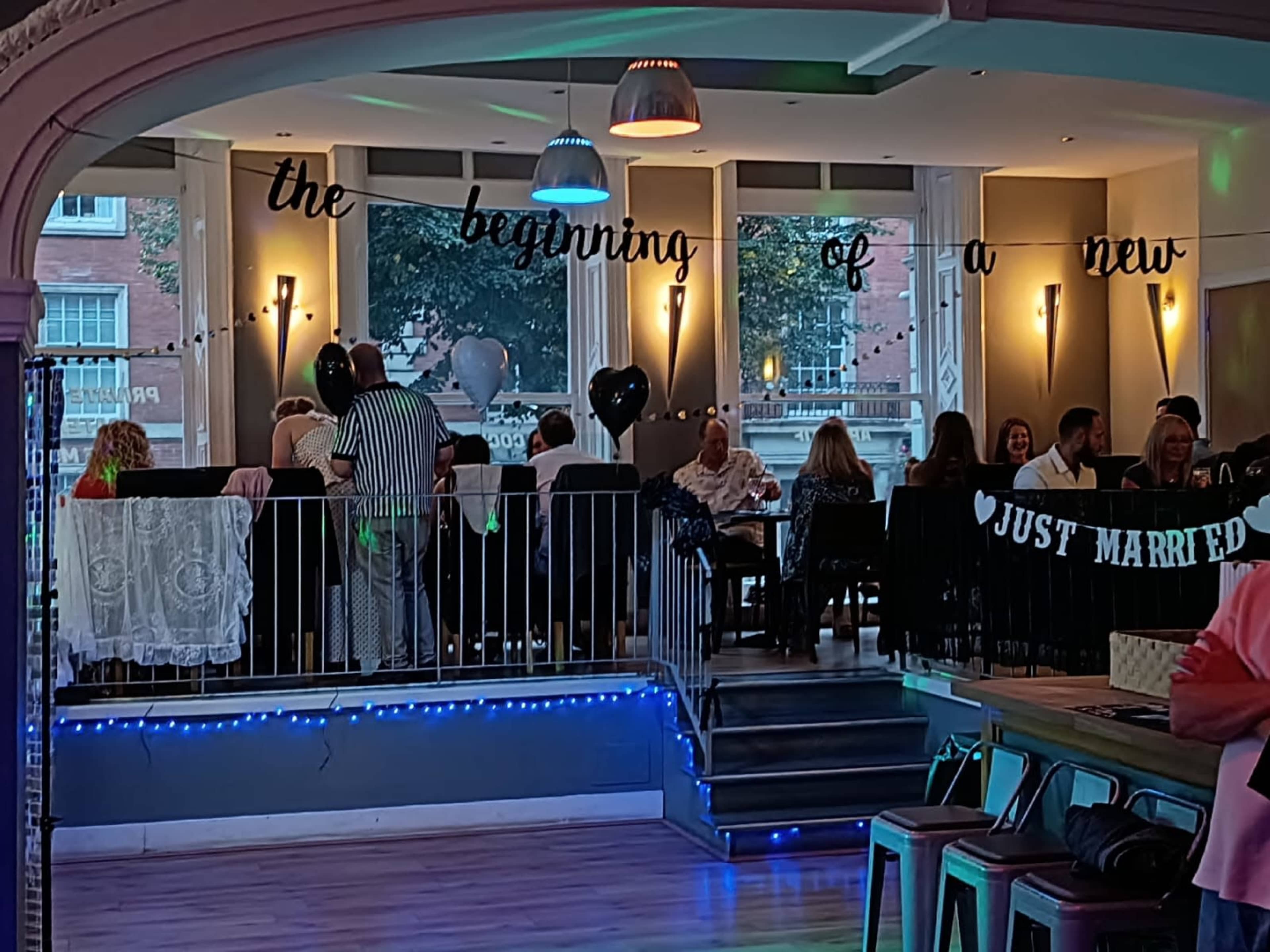 A celebratory gathering is taking place in a brightly lit venue, with guests seated at tables adorned with decorations and a banner that reads "Just Married."