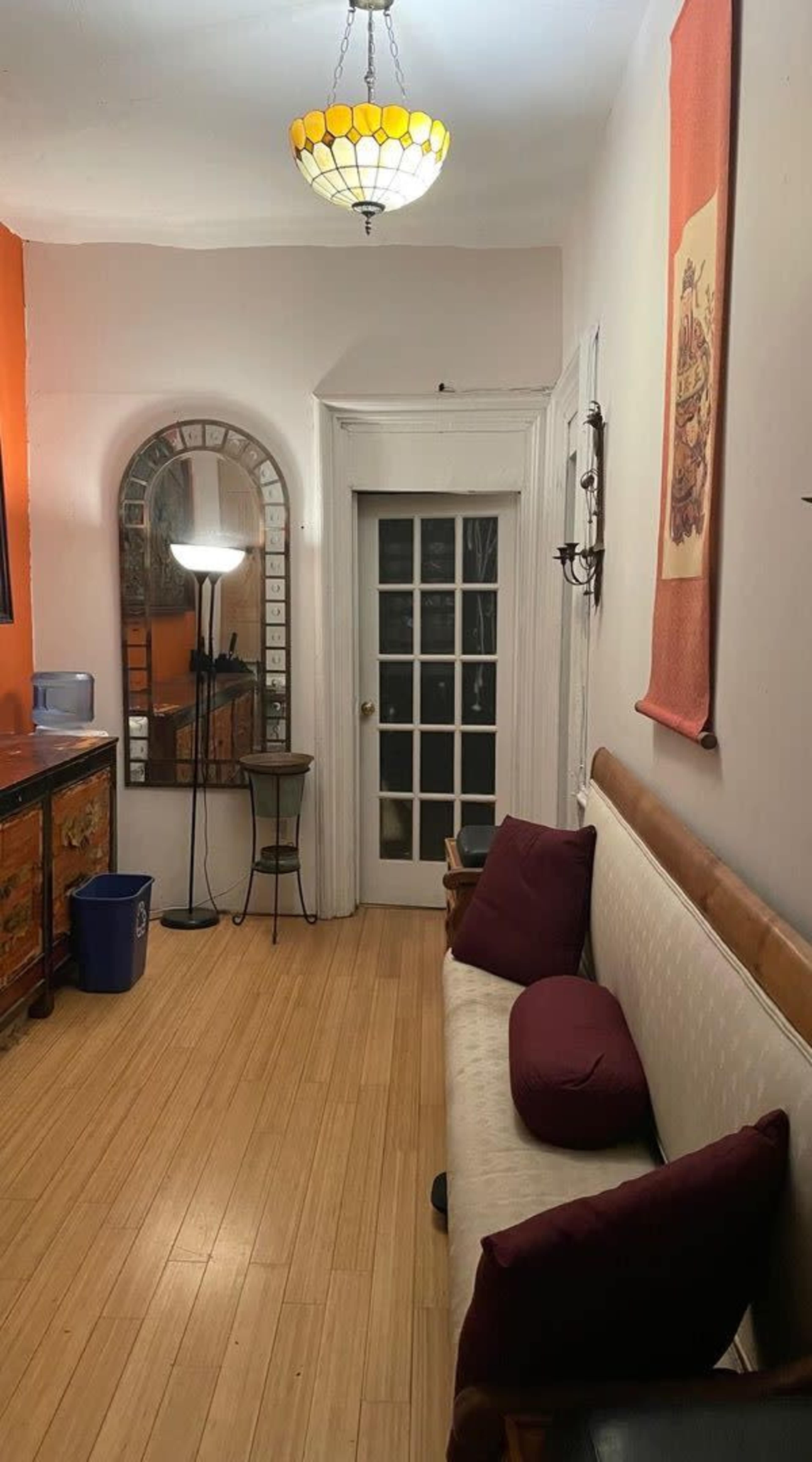 The image shows a small entryway with a wooden bench, a mirror, a modern floor lamp, and a door with glass panels, illuminated by a hanging light fixture.