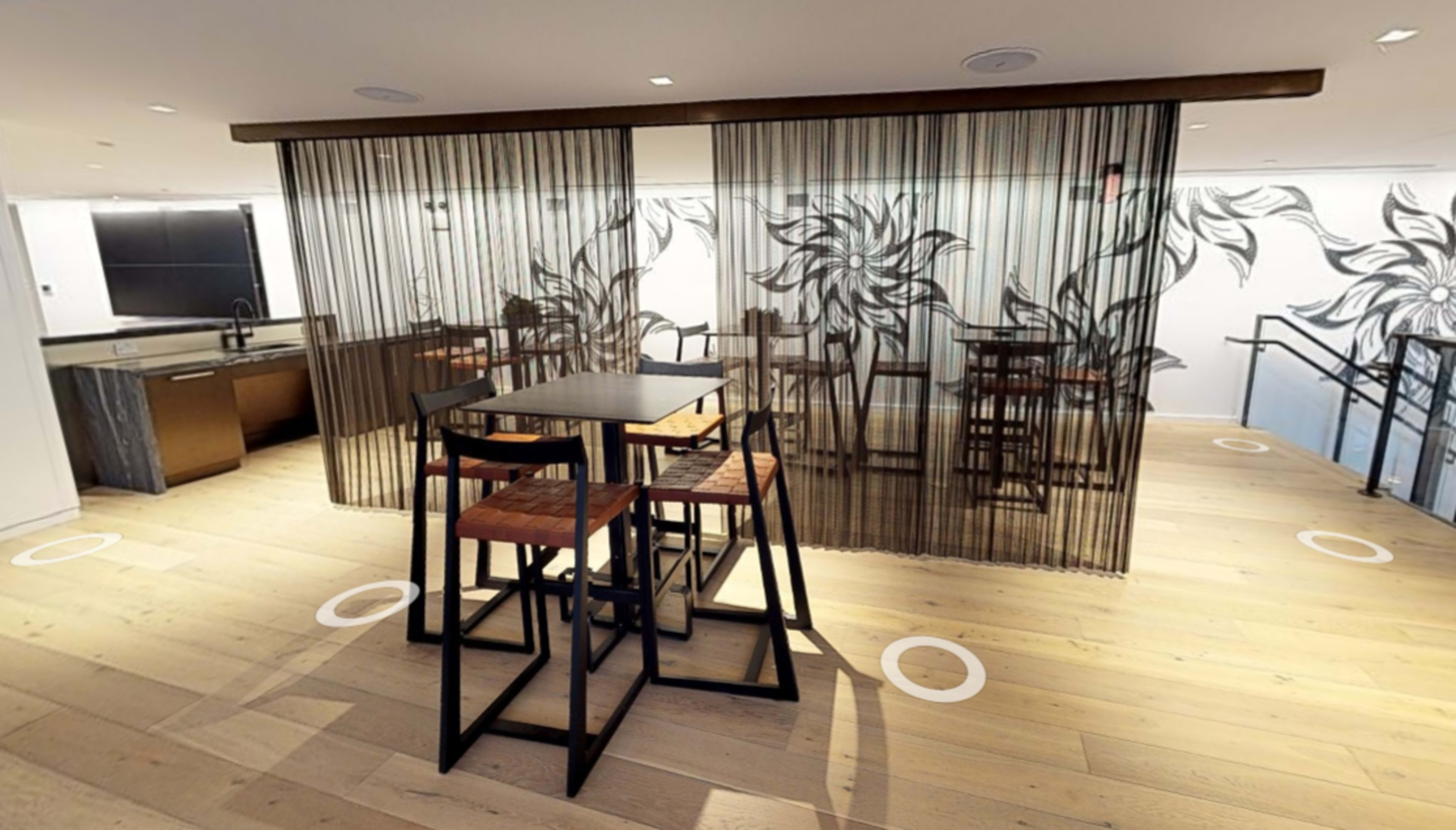 A modern dining area with a black and white floral motif curtain, a table with four stools, and a kitchen area in the background.