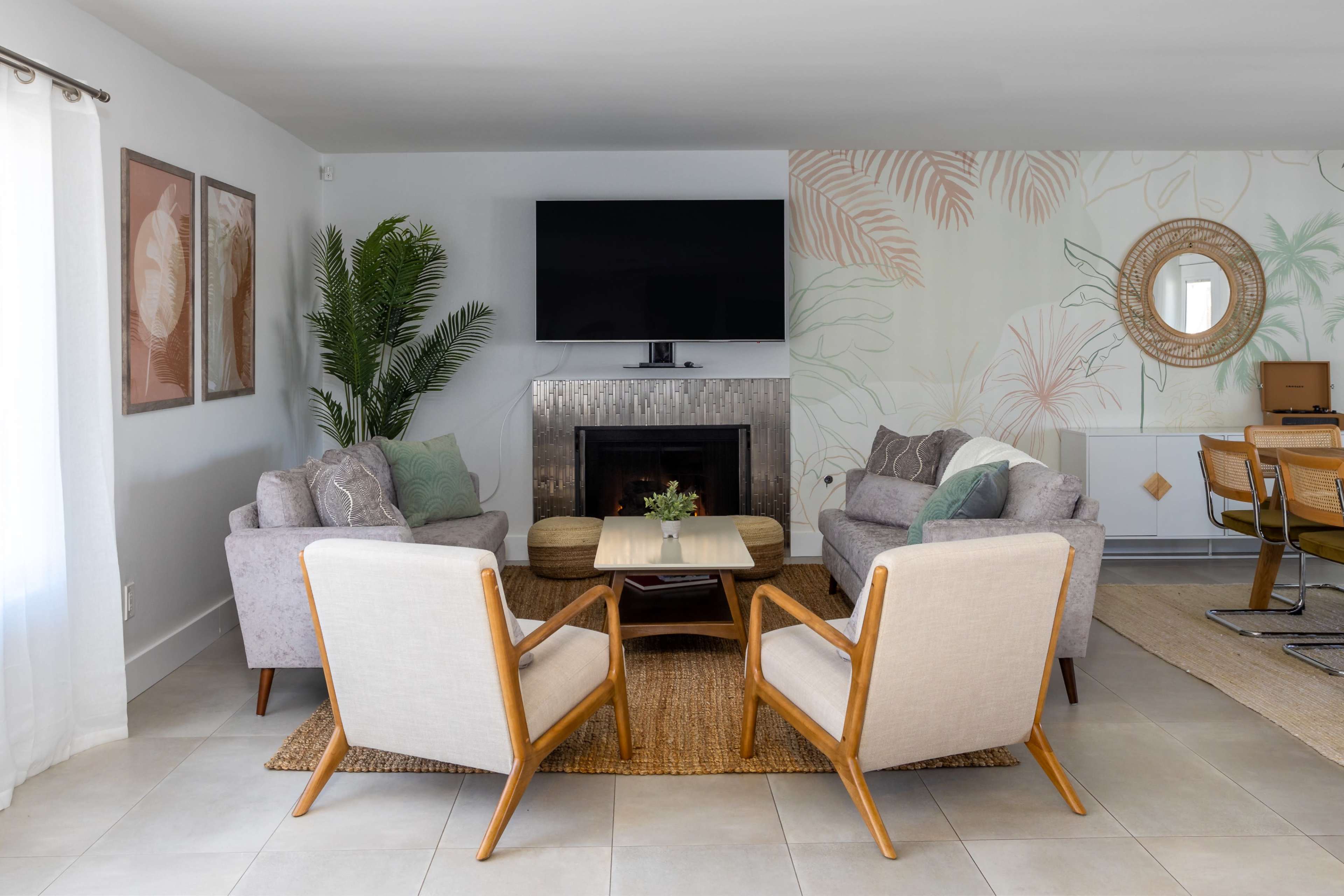 A living room features two gray sofas and two light-colored armchairs arranged around a wooden coffee table, with a large television mounted above a fireplace and decorative plants in the corner.