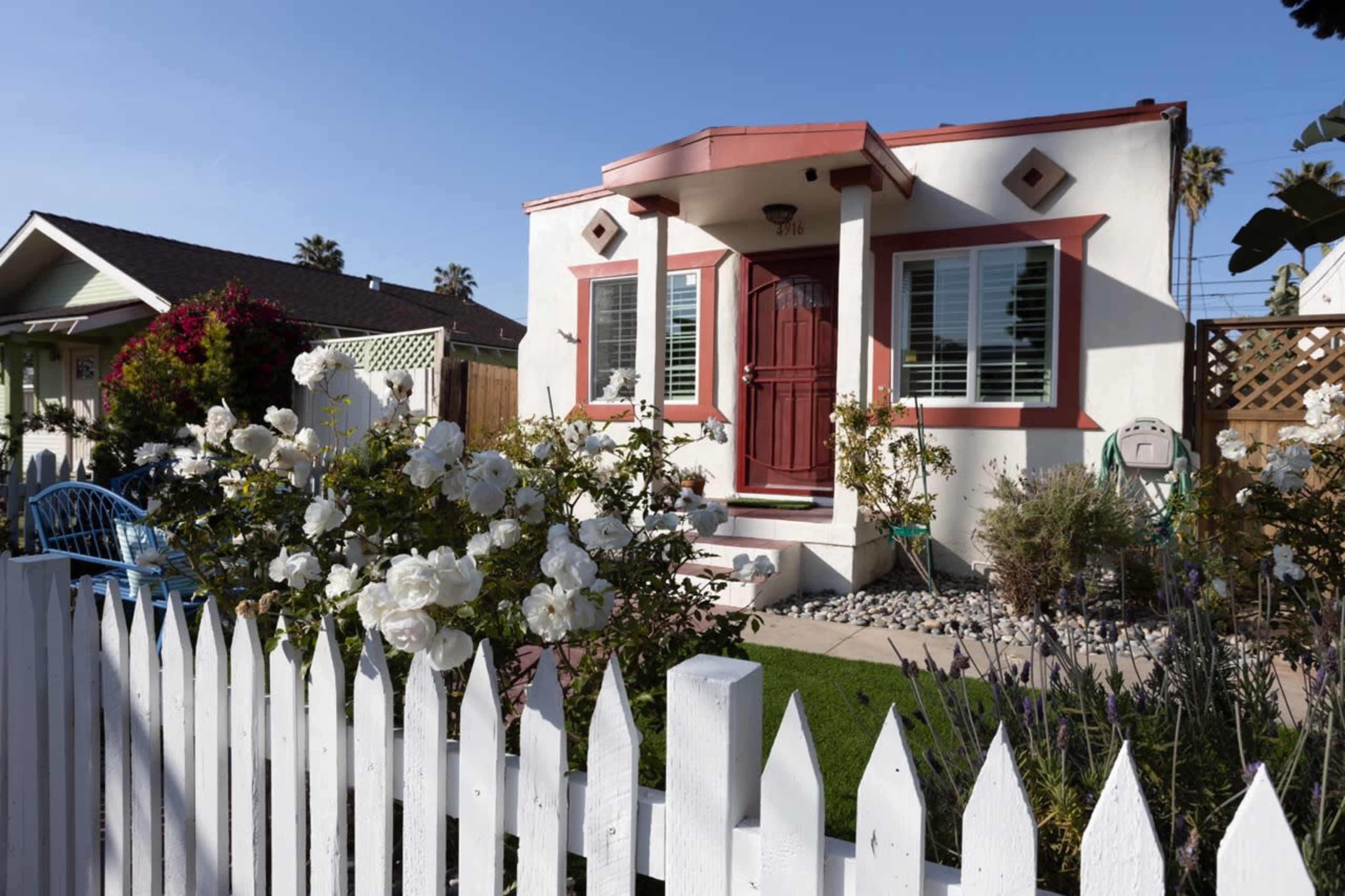A small, white house with red trim features a front porch and a neatly kept garden surrounded by a white picket fence.