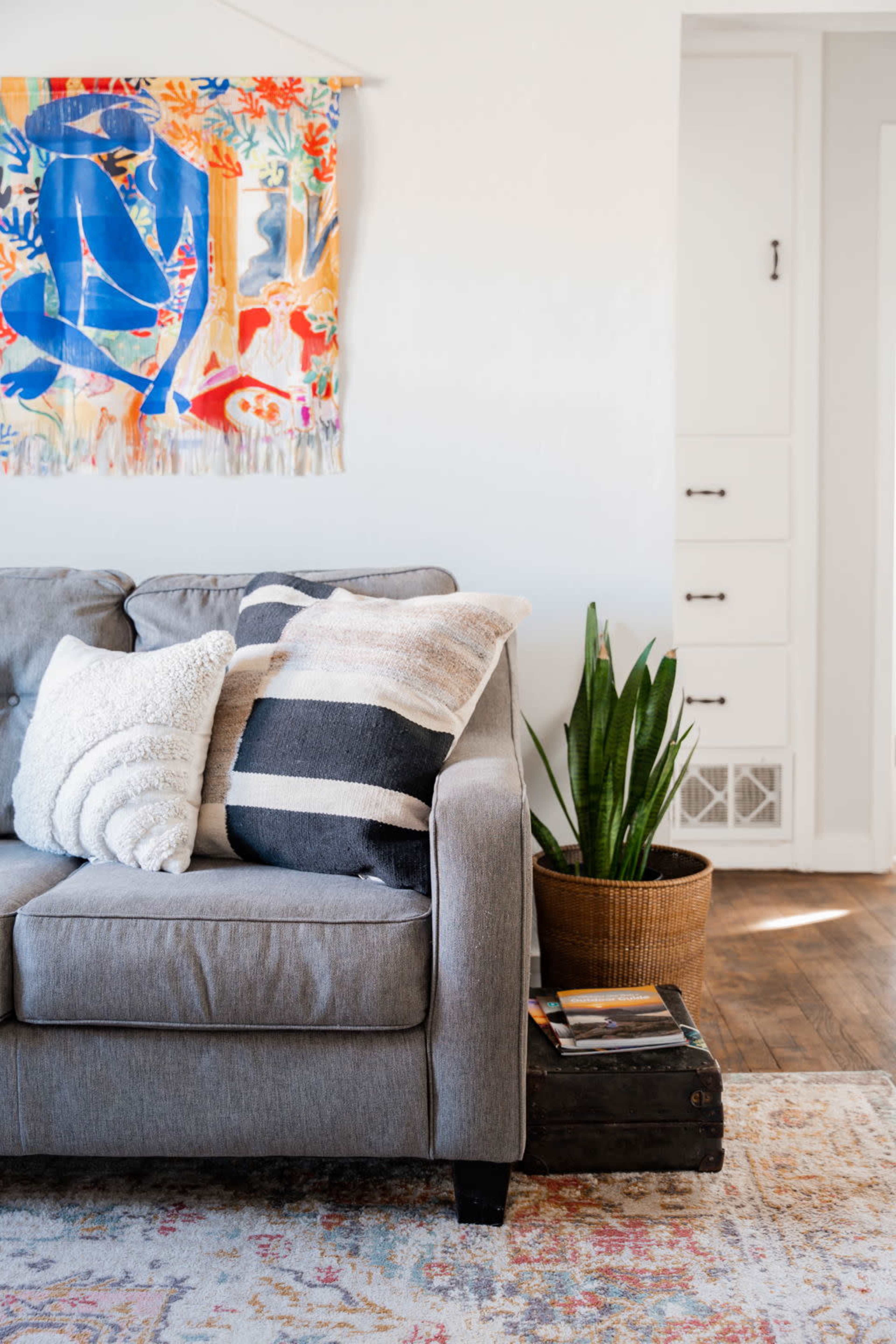 A gray sofa adorned with various cushions is positioned next to a potted plant and a stack of books on a wooden floor, while a colorful wall hanging decorates the background.