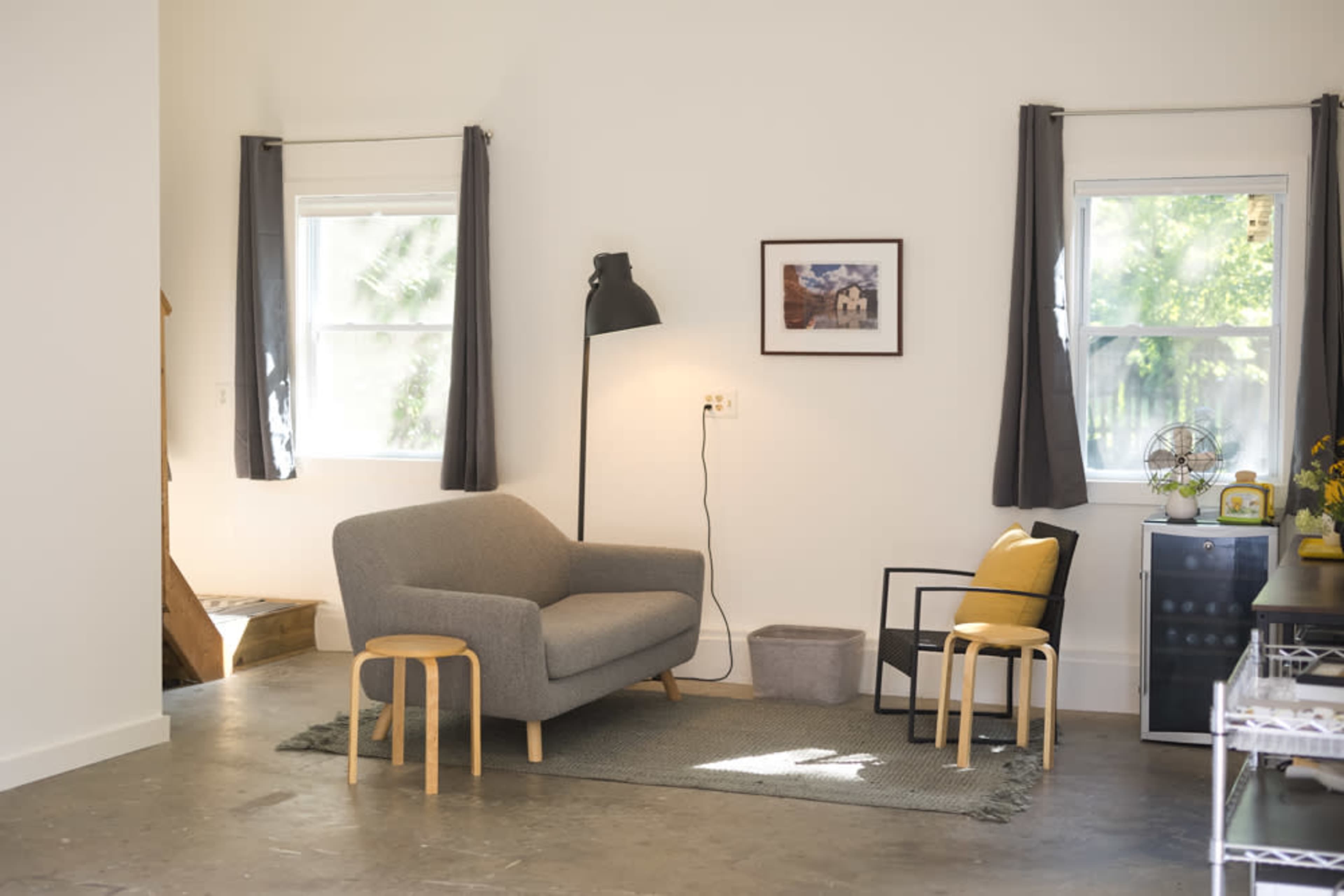 The image shows a minimalist living space featuring a gray sofa, a black chair with a yellow cushion, and a lamp beside a small table on a rug.