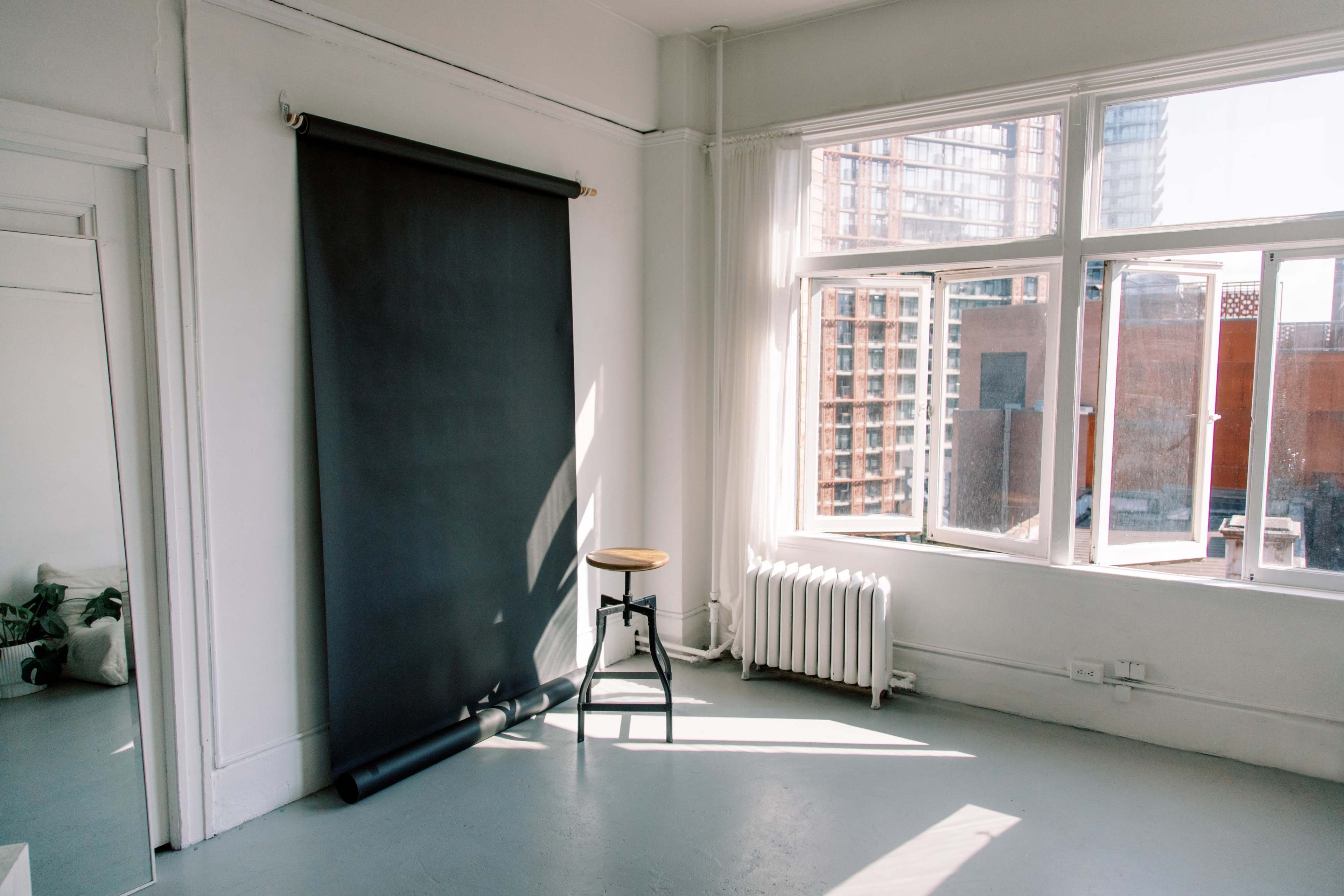 The image shows a minimalist room with a black backdrop, a stool, and large windows allowing natural light to enter.