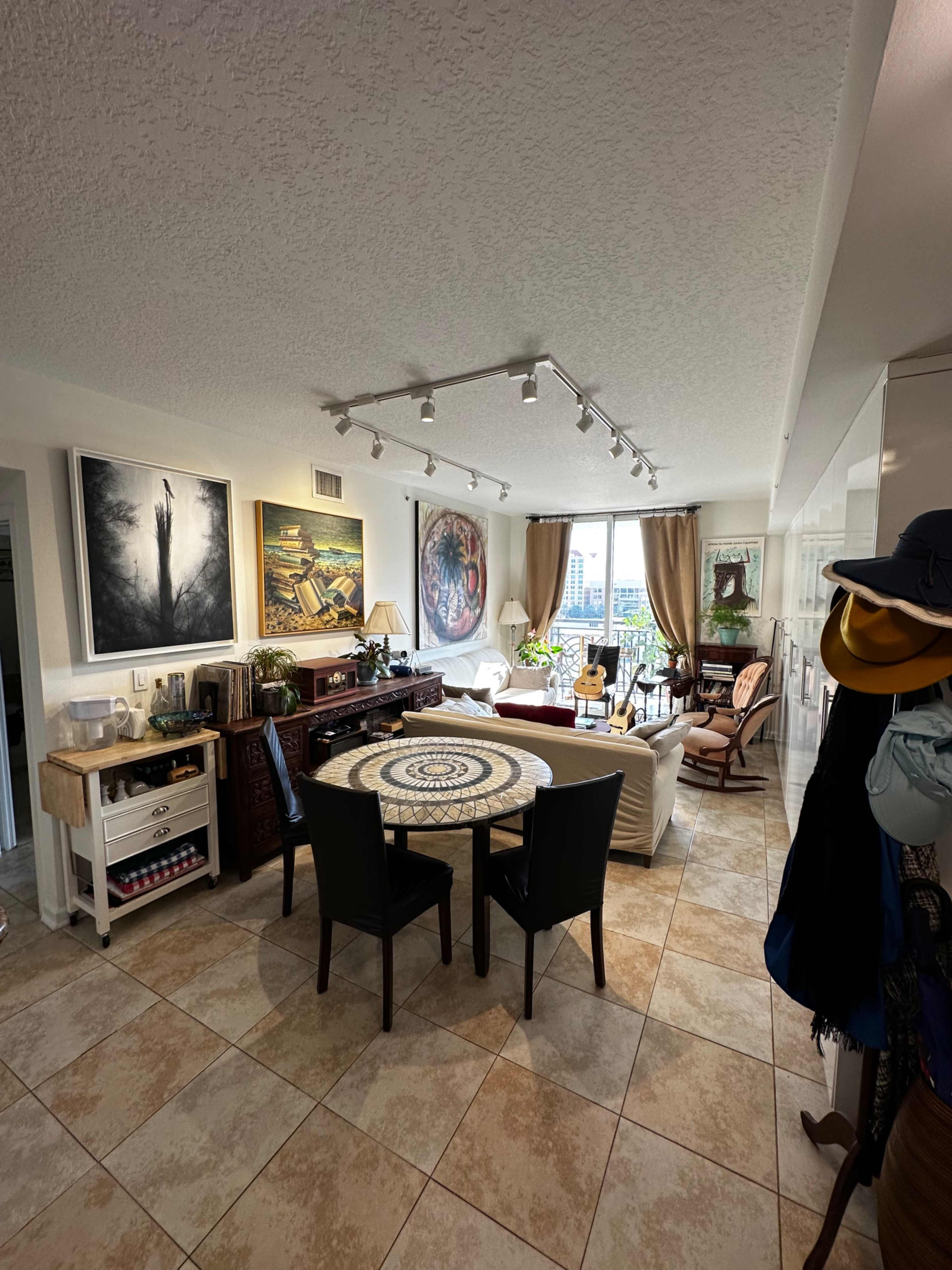 A well-lit living space features a dining area with a round table, a couch by a large window, and artwork on the walls.