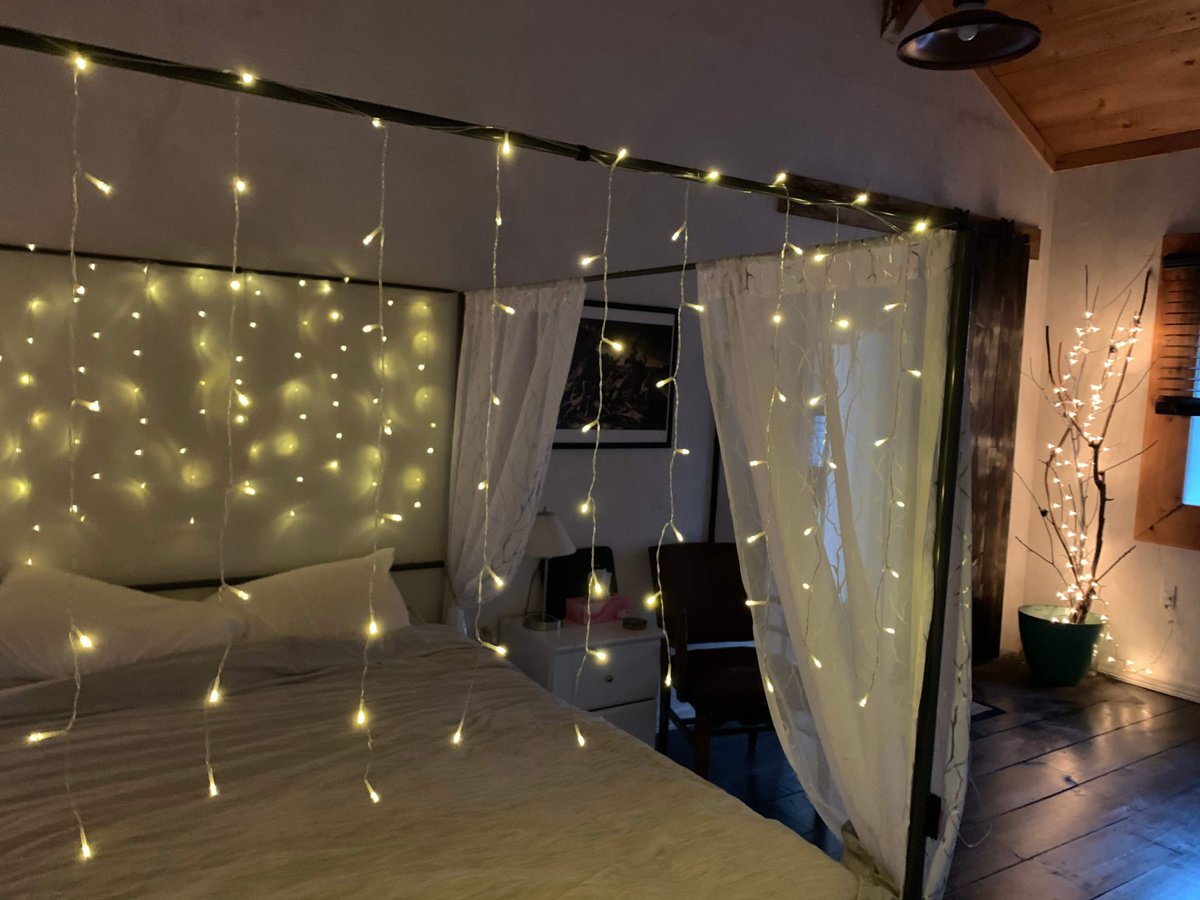 A bedroom features a bed draped with white curtains and adorned with string lights, alongside a potted plant in the corner.