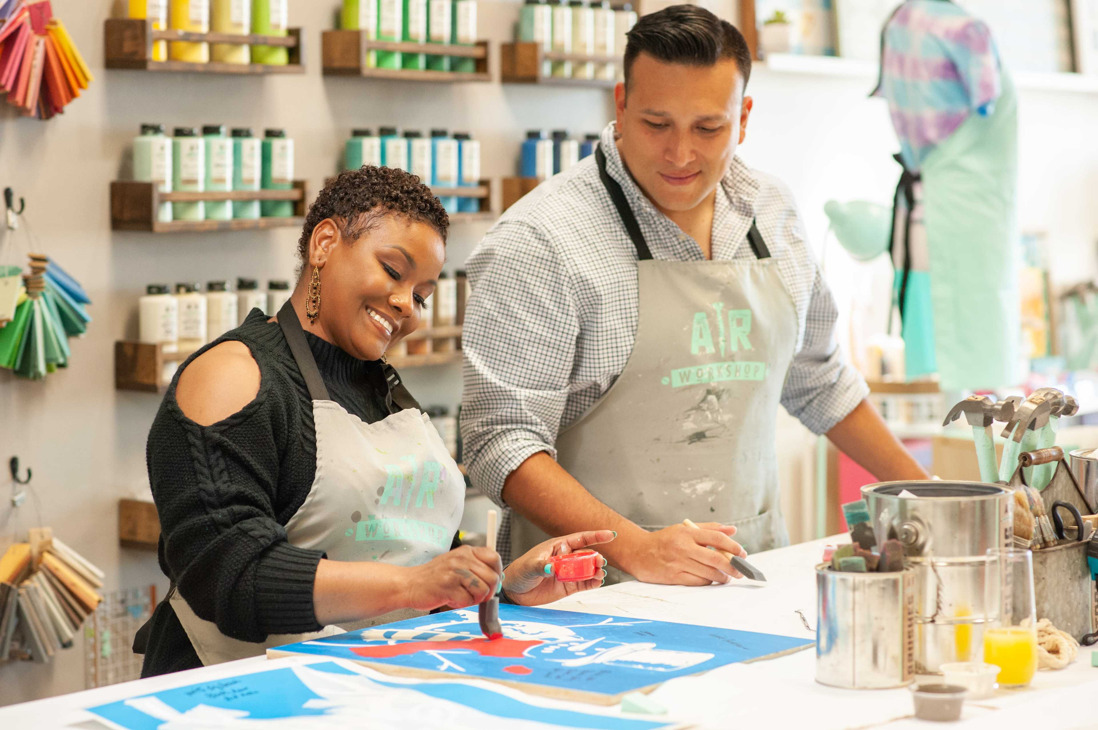 Two artists are working on a colorful painting project at a table filled with art supplies in a well-lit studio.
