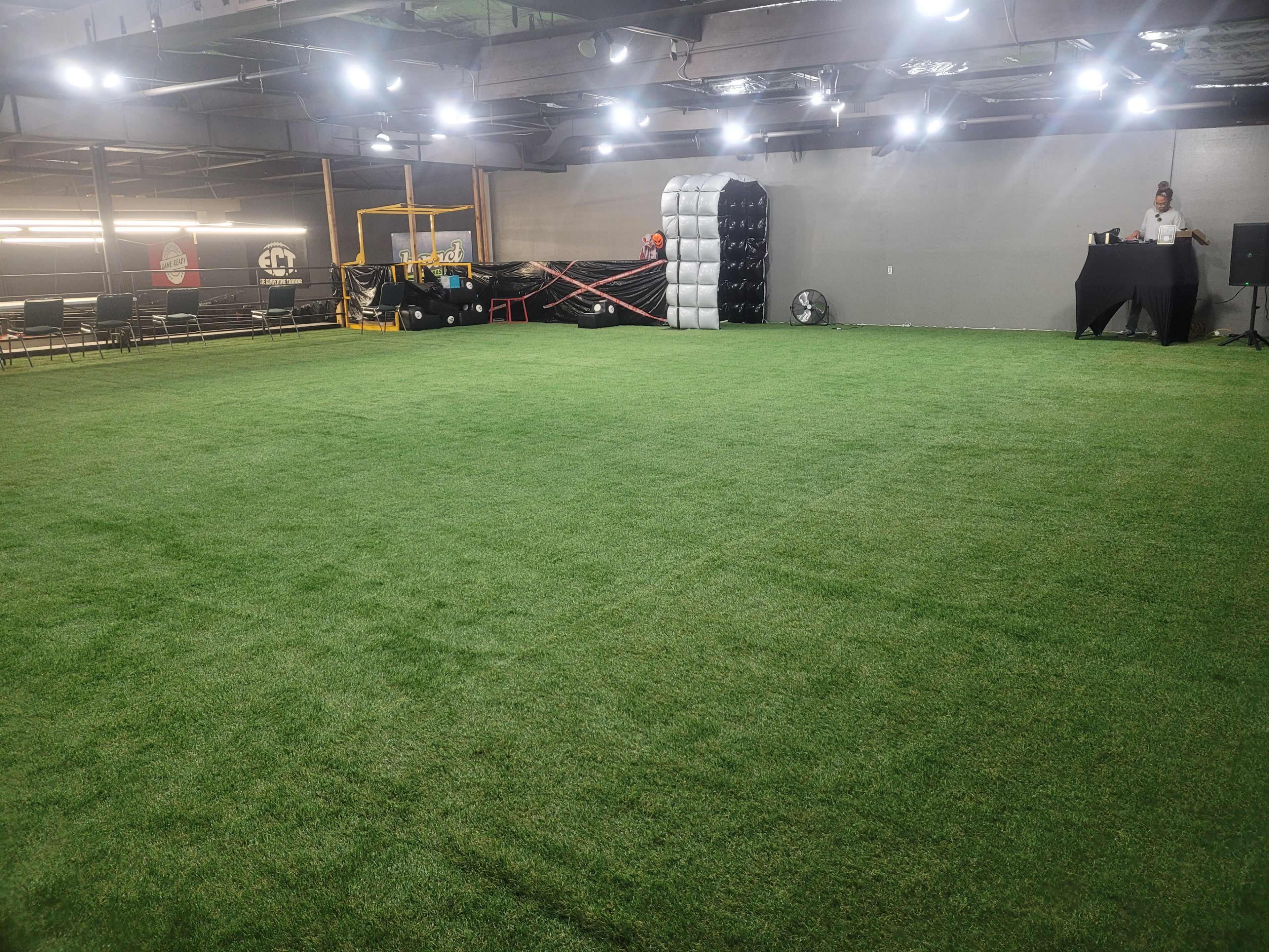 The image shows a spacious indoor area with green artificial turf, empty chairs arranged along the sides, and a black table set up in one corner.
