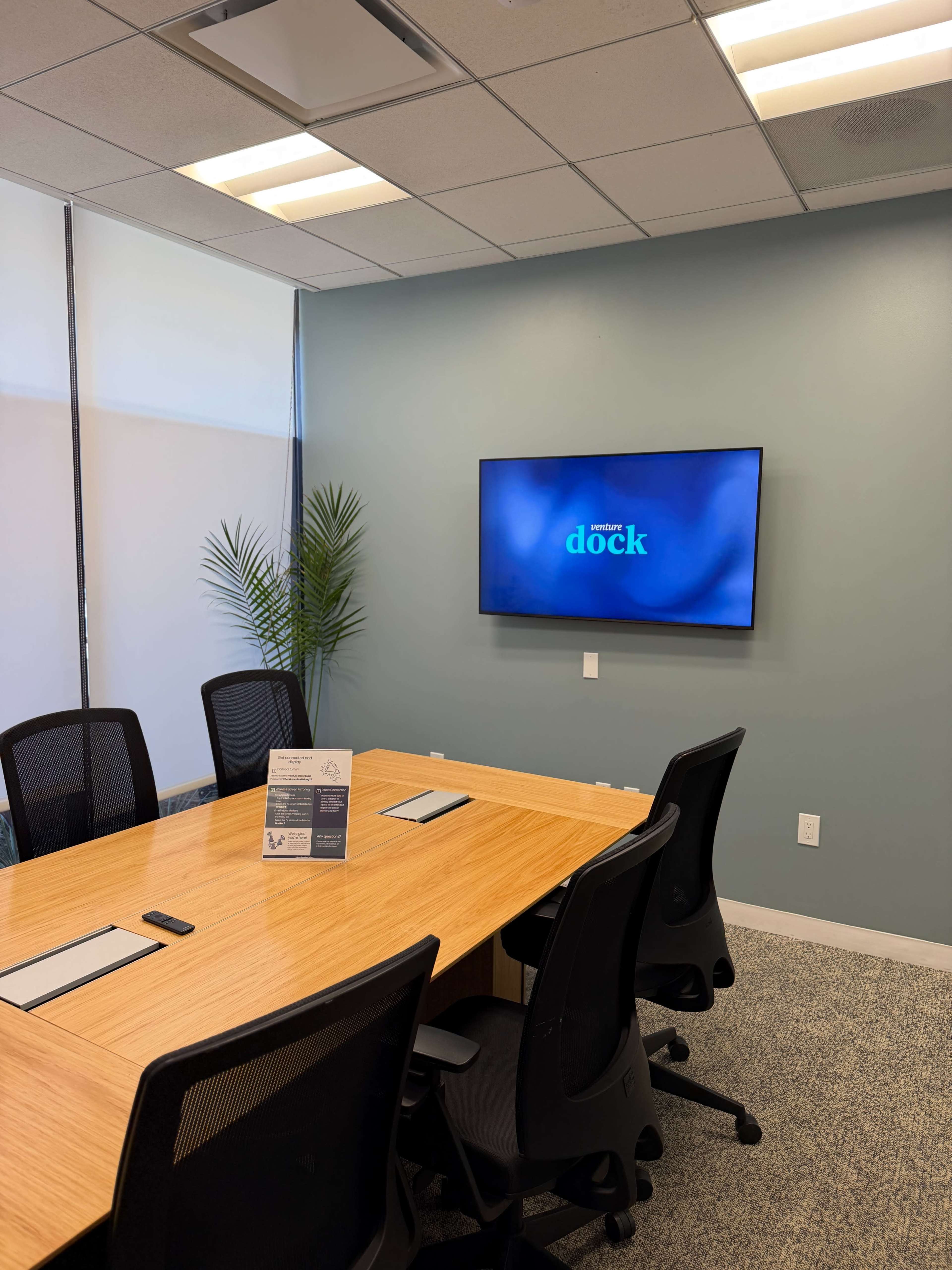 The image shows a modern conference room featuring a large wooden table, black ergonomic chairs, a wall-mounted television displaying the word "dock," and a potted plant in the corner.