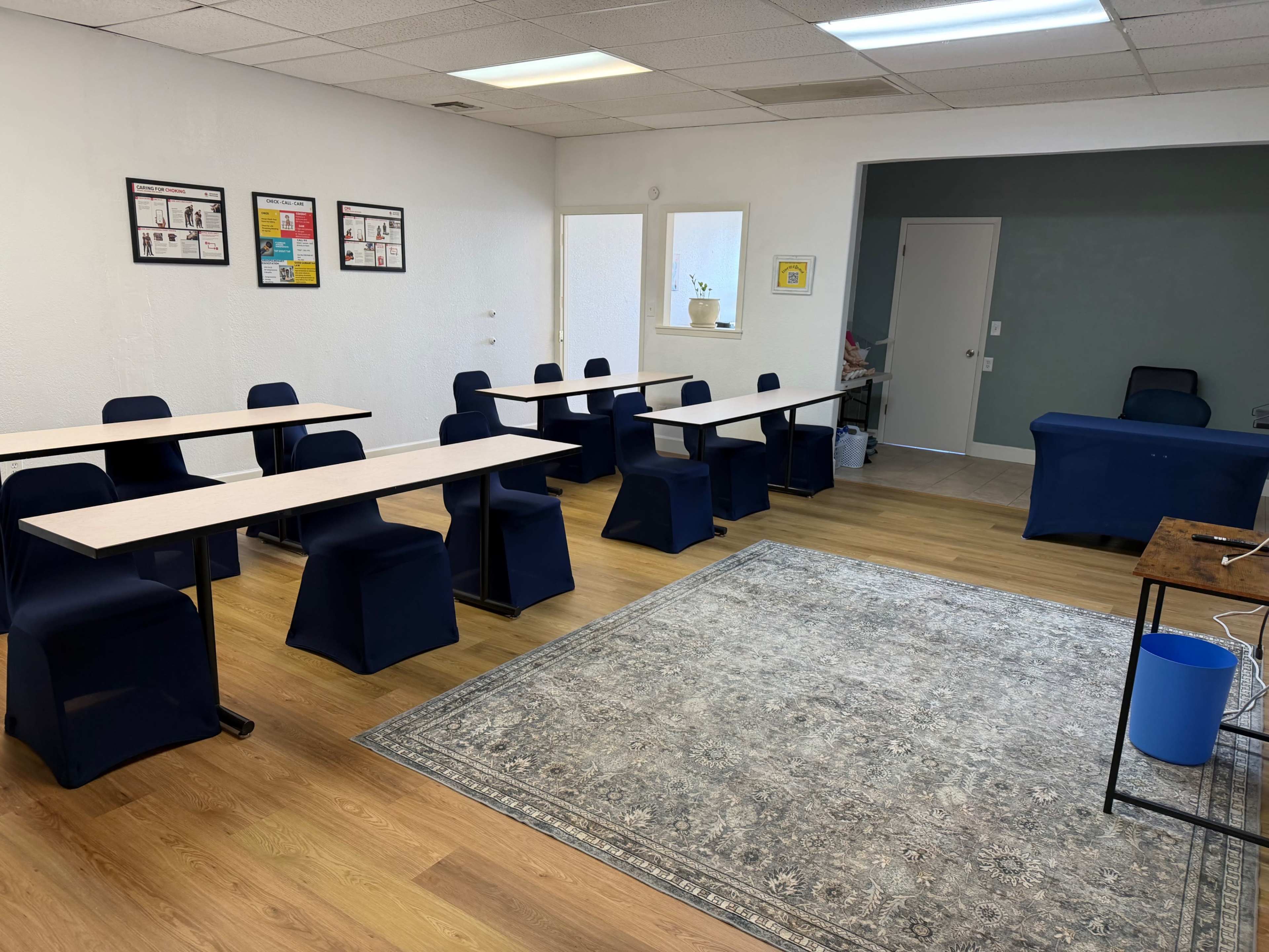 The image shows a classroom or meeting space with several tables and chairs arranged neatly, a rug on the floor, and a reception desk at the front.