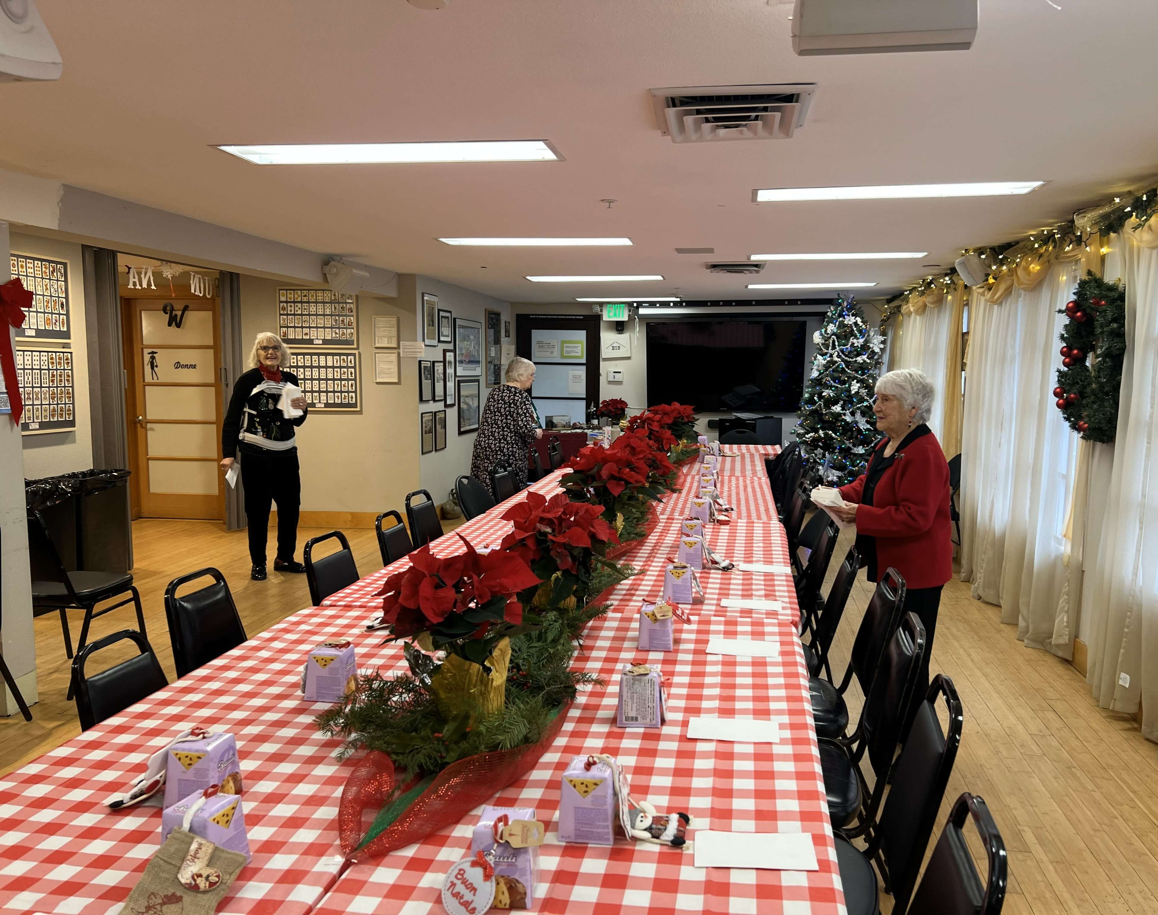 A long table with a red and white checkered tablecloth is set for a holiday gathering, flanked by chairs and decorated with poinsettias and festive centerpieces, while two women prepare the space.