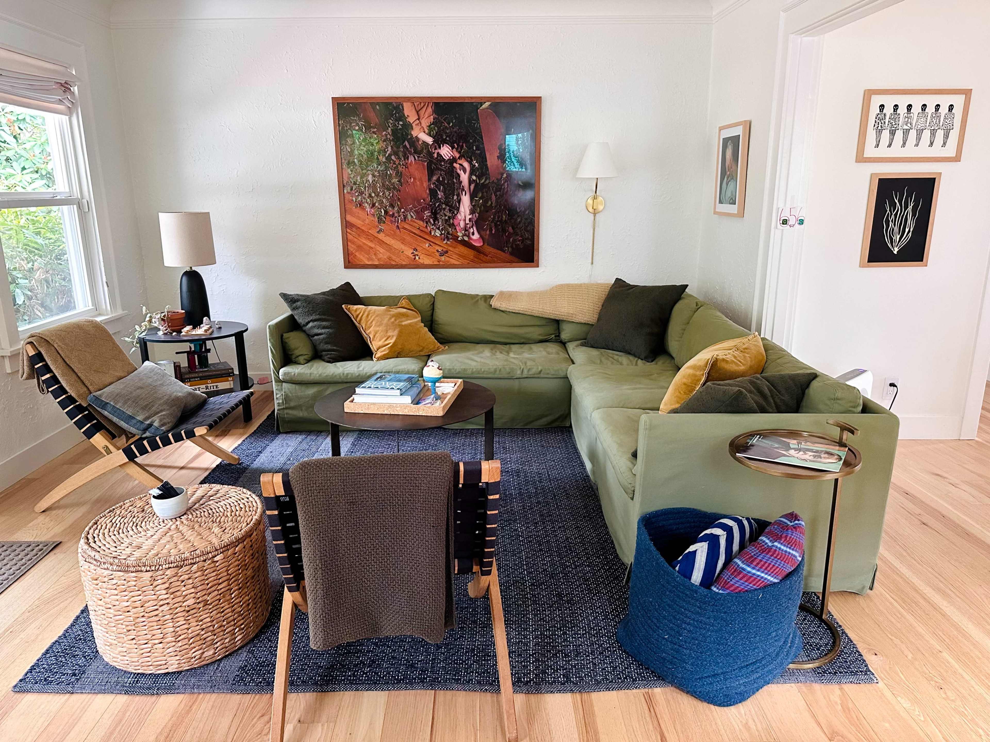 The image shows a cozy living room with a green sectional sofa, a black table in the center, a woven basket as a side table, and various decorative accents on the walls.