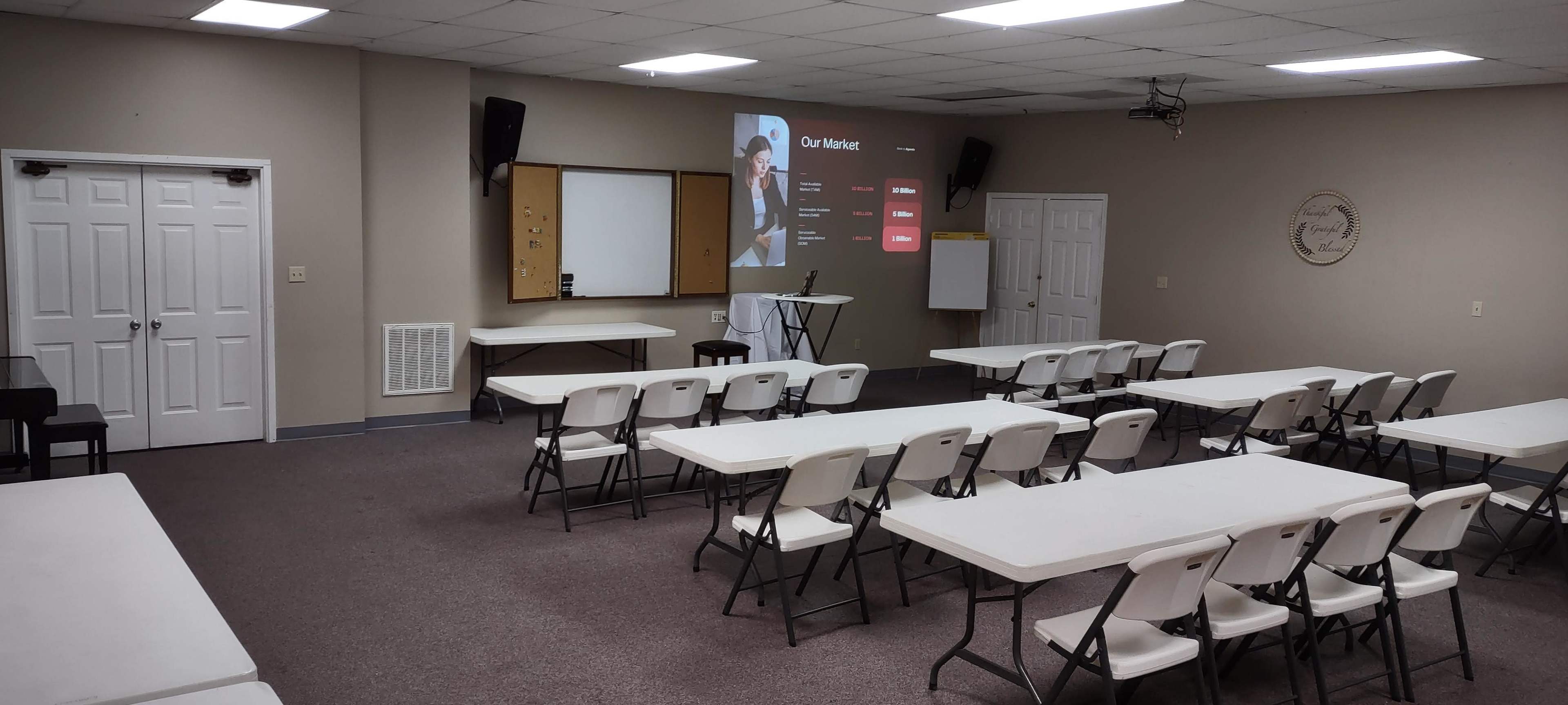 A classroom setting with rows of white tables and chairs, a projector screen displaying a presentation, and a black stand in the front.