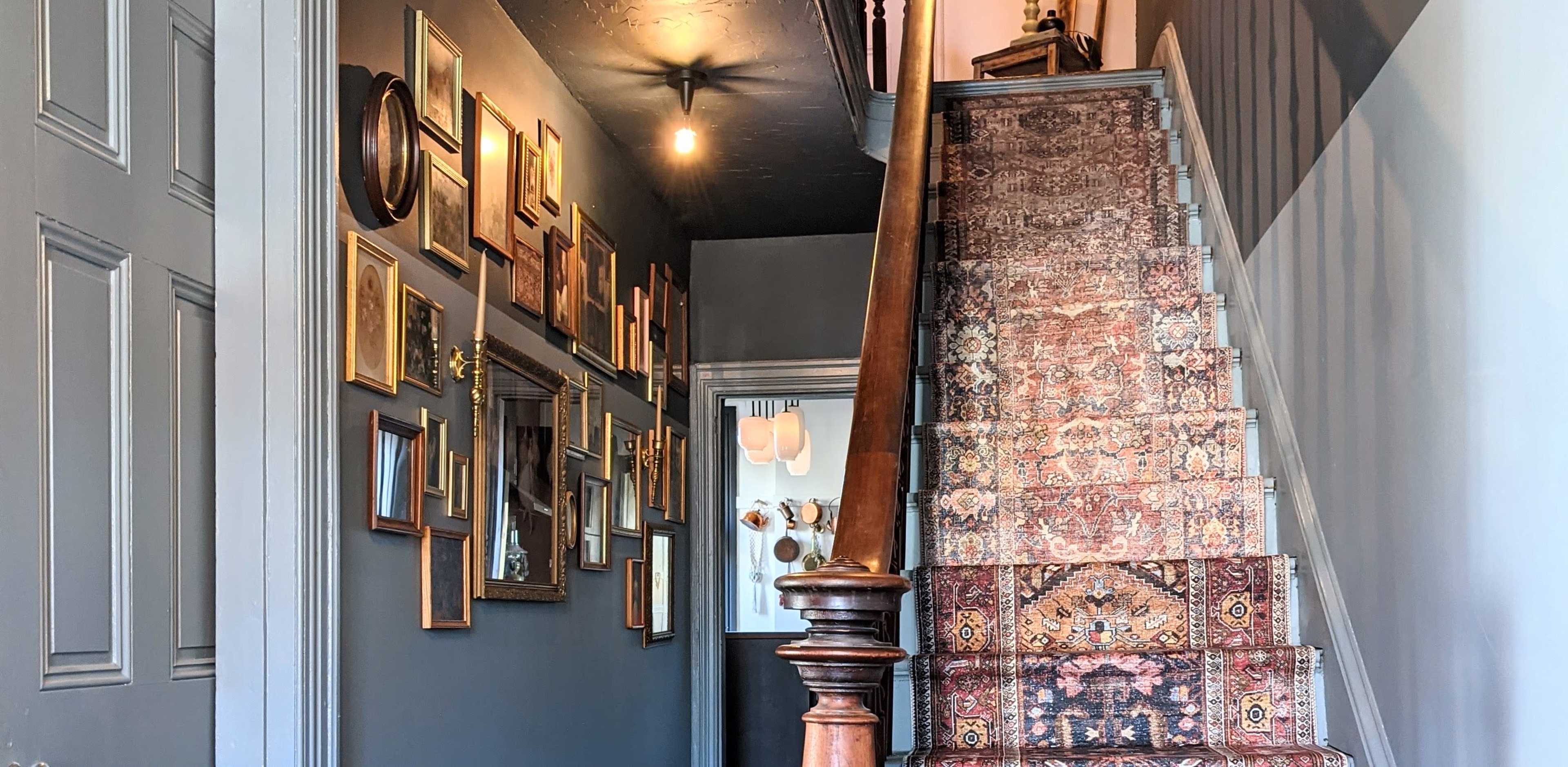 A staircase with a patterned rug, flanked by a wall decorated with various framed pictures, leading up to a dimly lit landing.