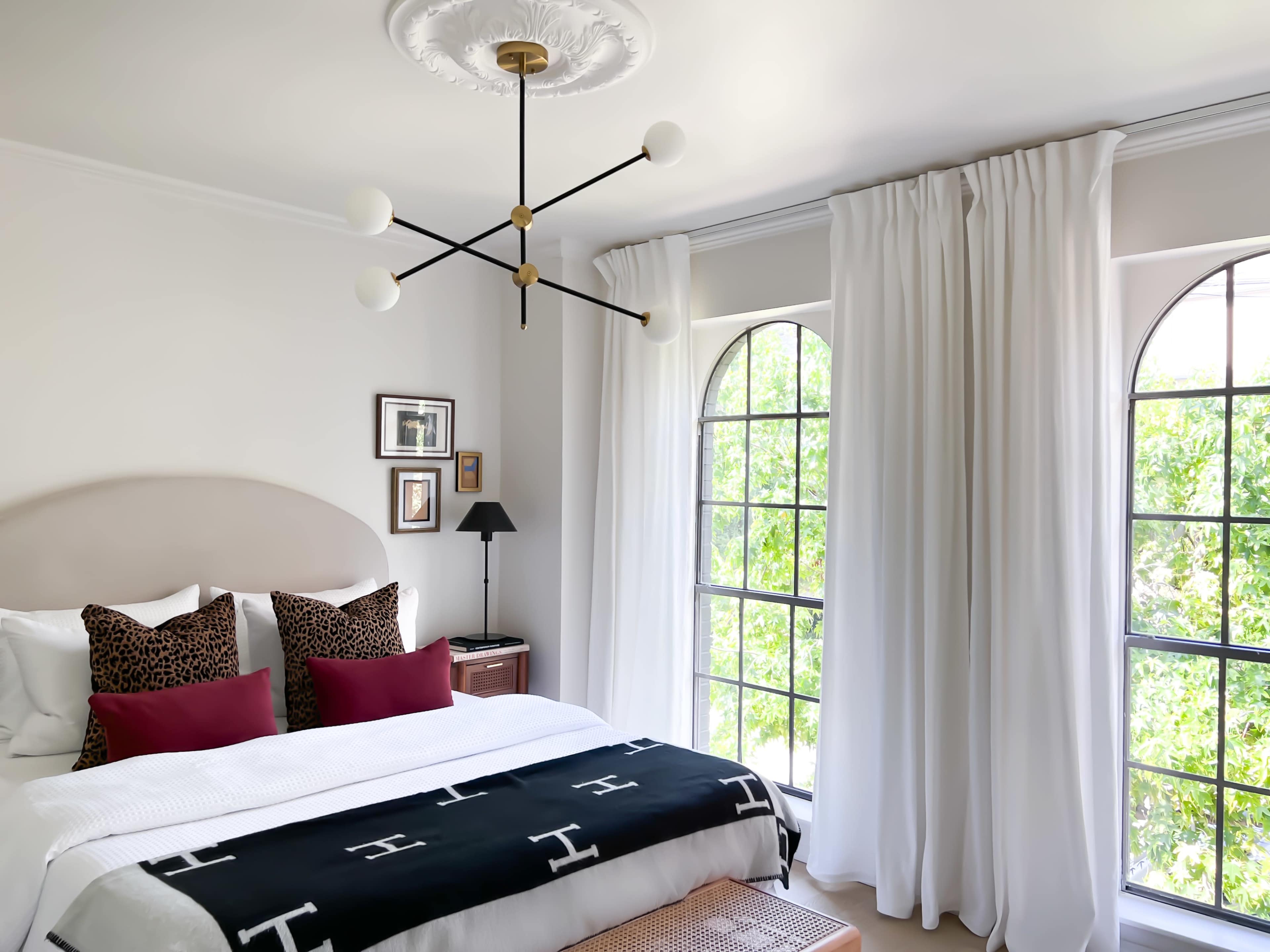 The bedroom features a bed with white linens and red accent pillows, flanked by large windows with white curtains and a modern light fixture overhead.