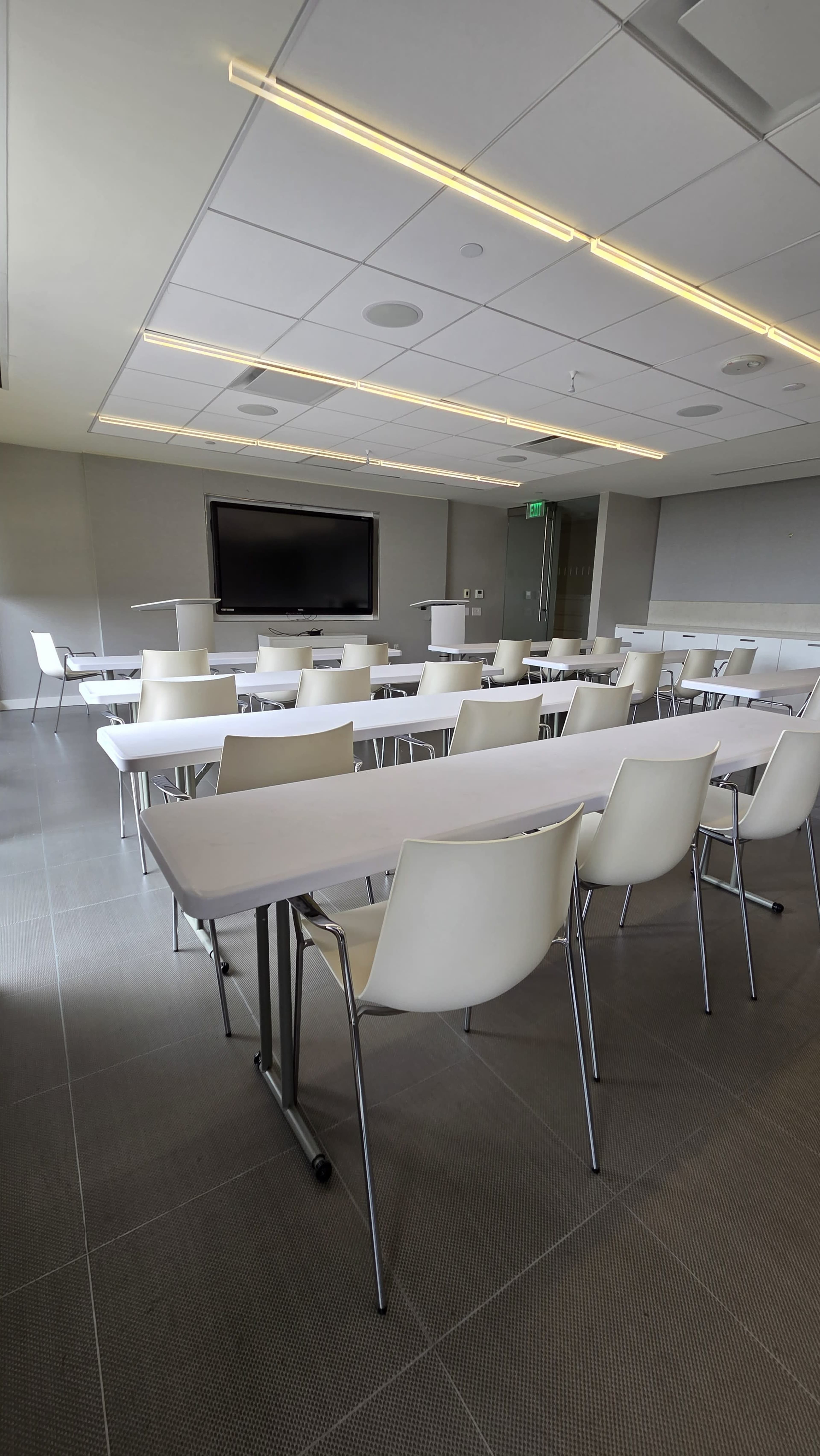 A modern classroom is set up with rows of white tables and chairs facing a wall-mounted screen.