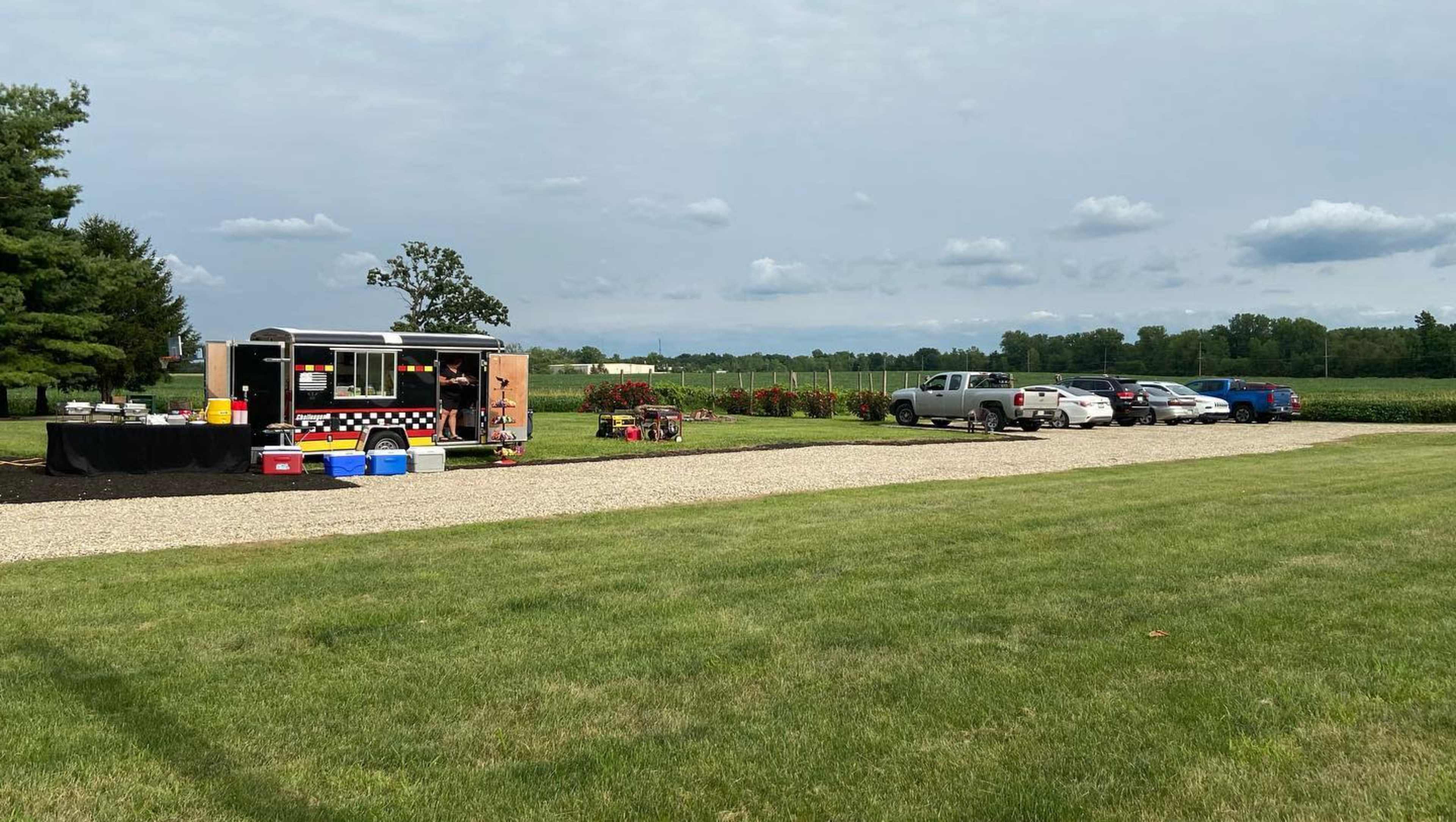 A food truck is parked beside a gravel lot where several vehicles are stationed, with fields and trees in the background.