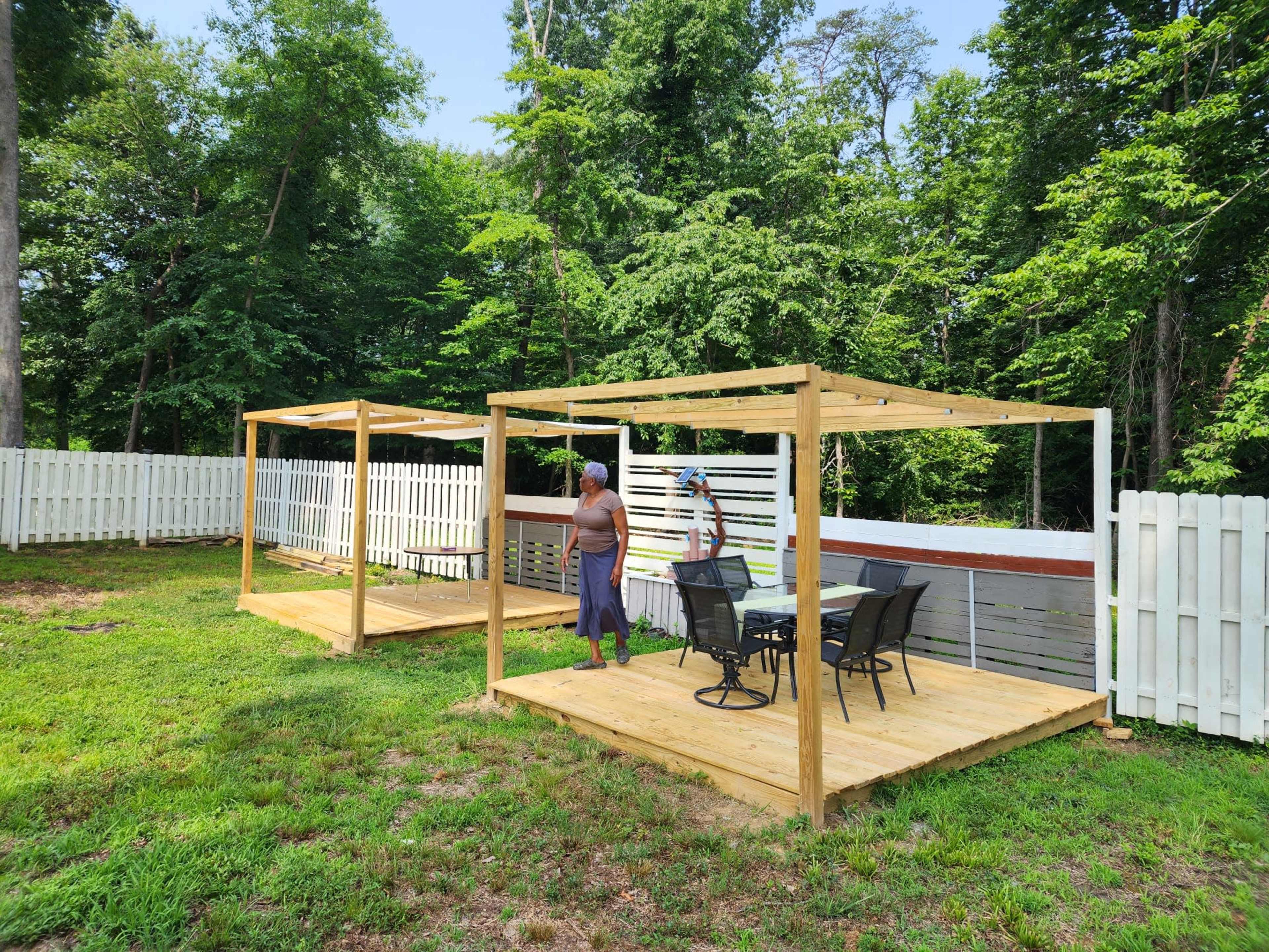Two wooden pergolas with chairs are set up on a grassy area surrounded by a white fence and trees.