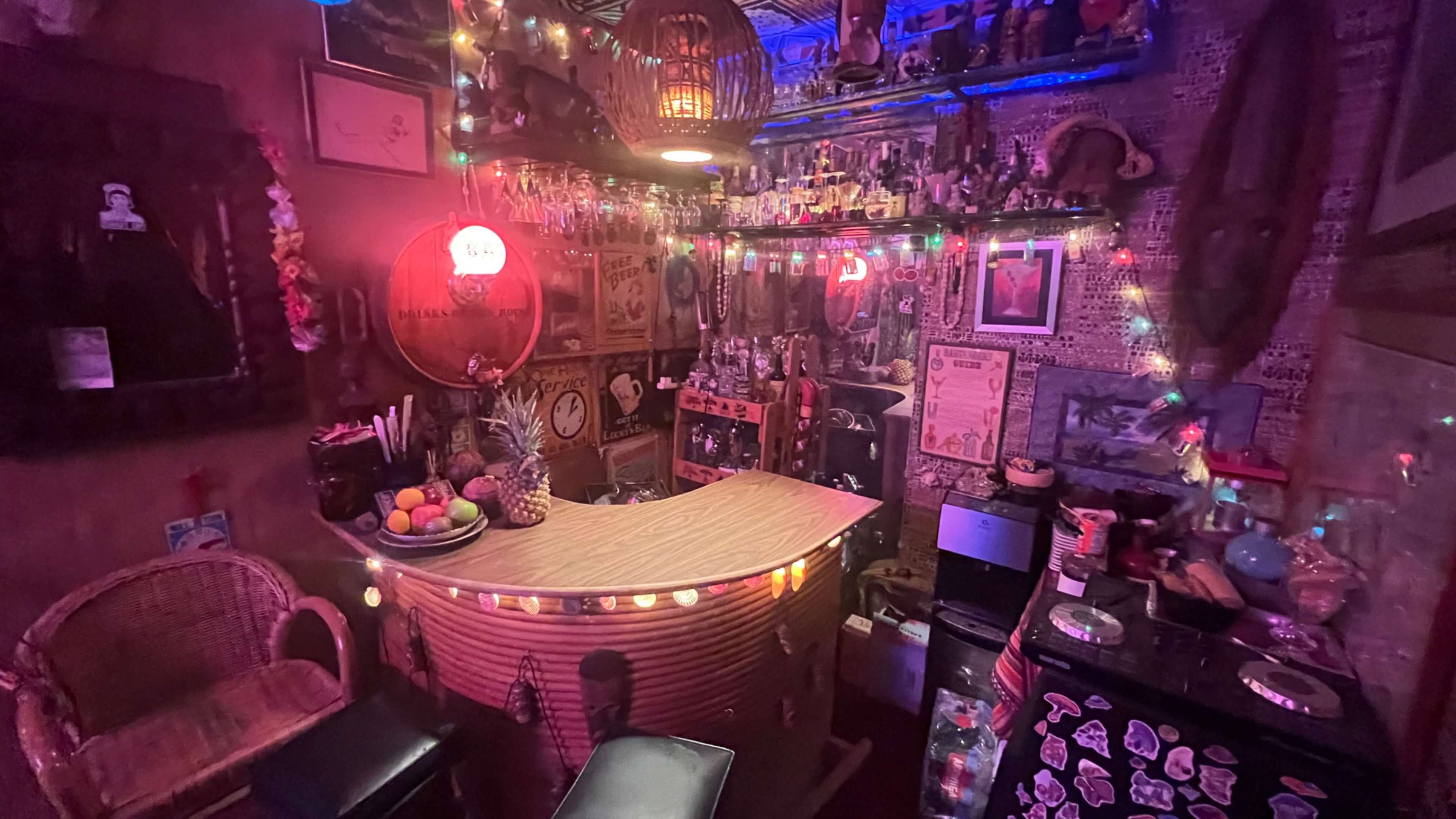 The image shows a dimly lit bar area adorned with colorful lights, tropical decorations, and various collectibles lining the walls.