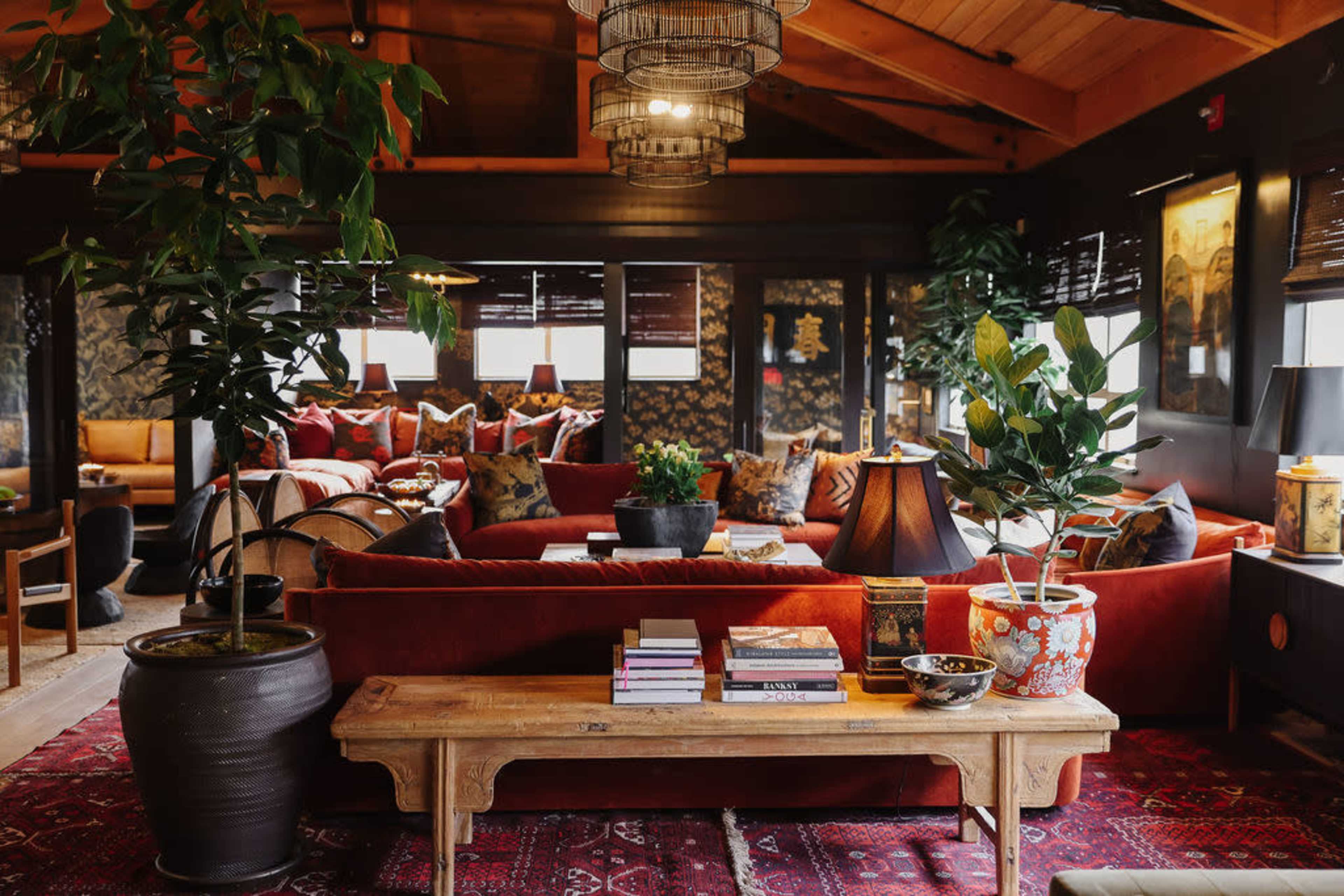 A cozy living room with a large red sectional sofa, various houseplants, and a wooden coffee table stacked with books.