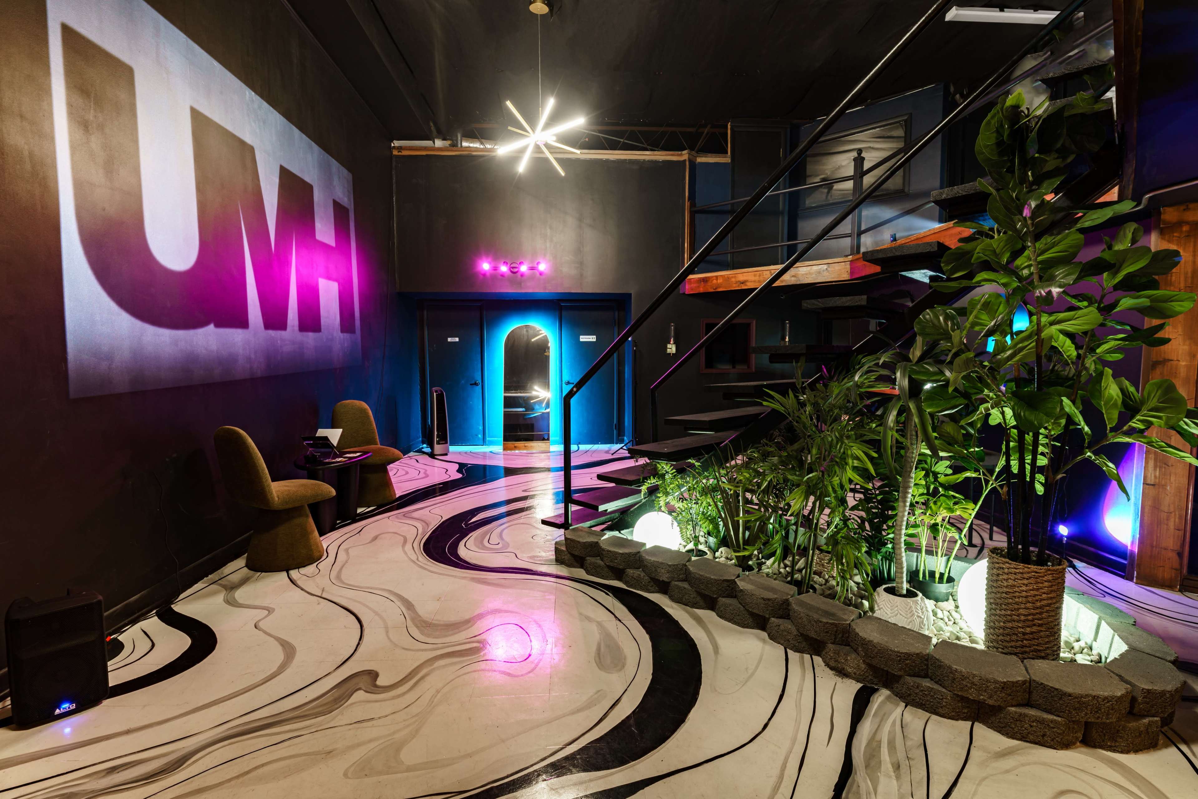 The image depicts a stylish interior space featuring a curved marble floor, modern seating, and illuminated plants, with a staircase leading to a second level.