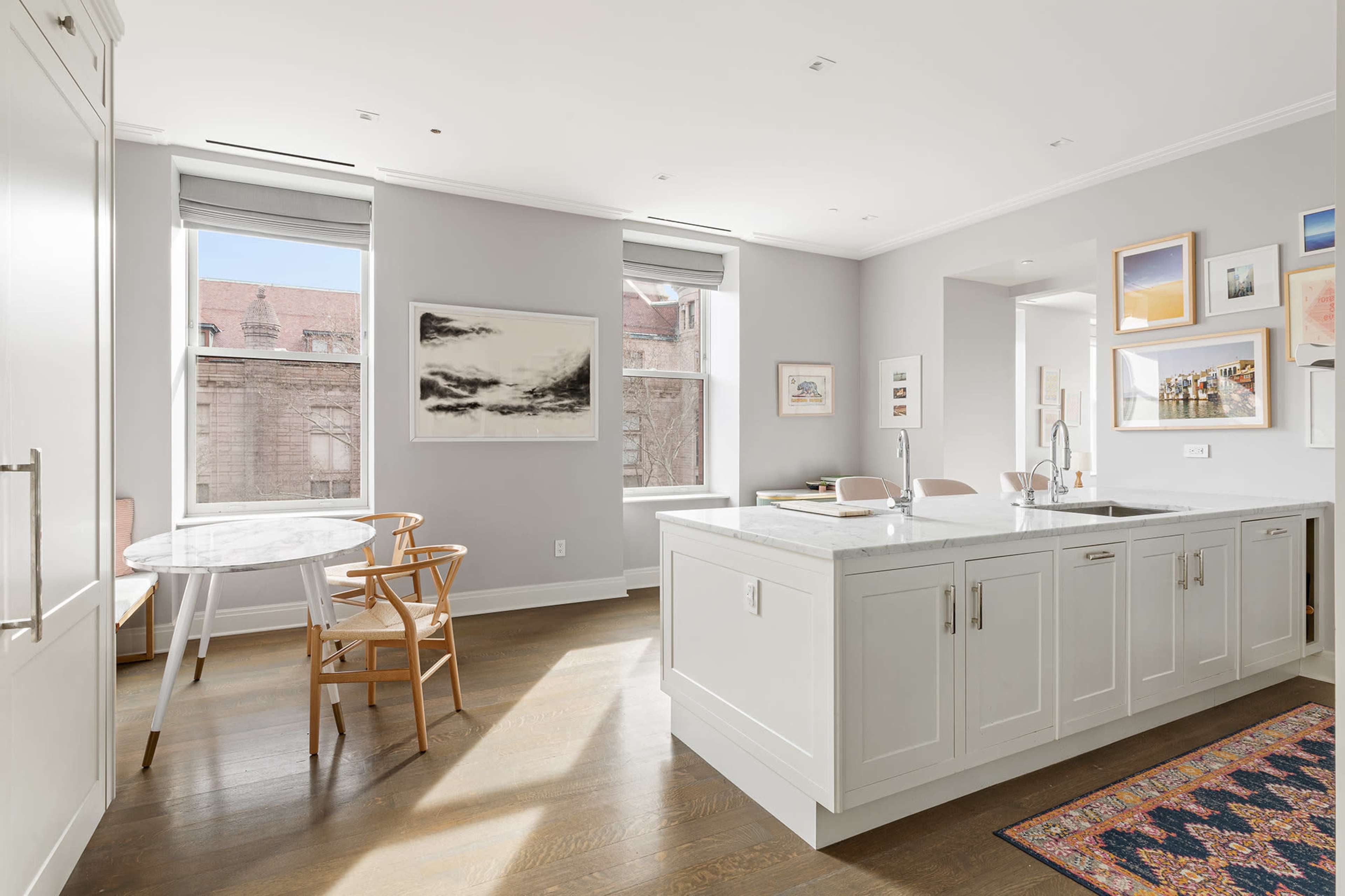 The image shows a modern kitchen and dining area with a marble-topped table, wooden chairs, and large windows allowing natural light.